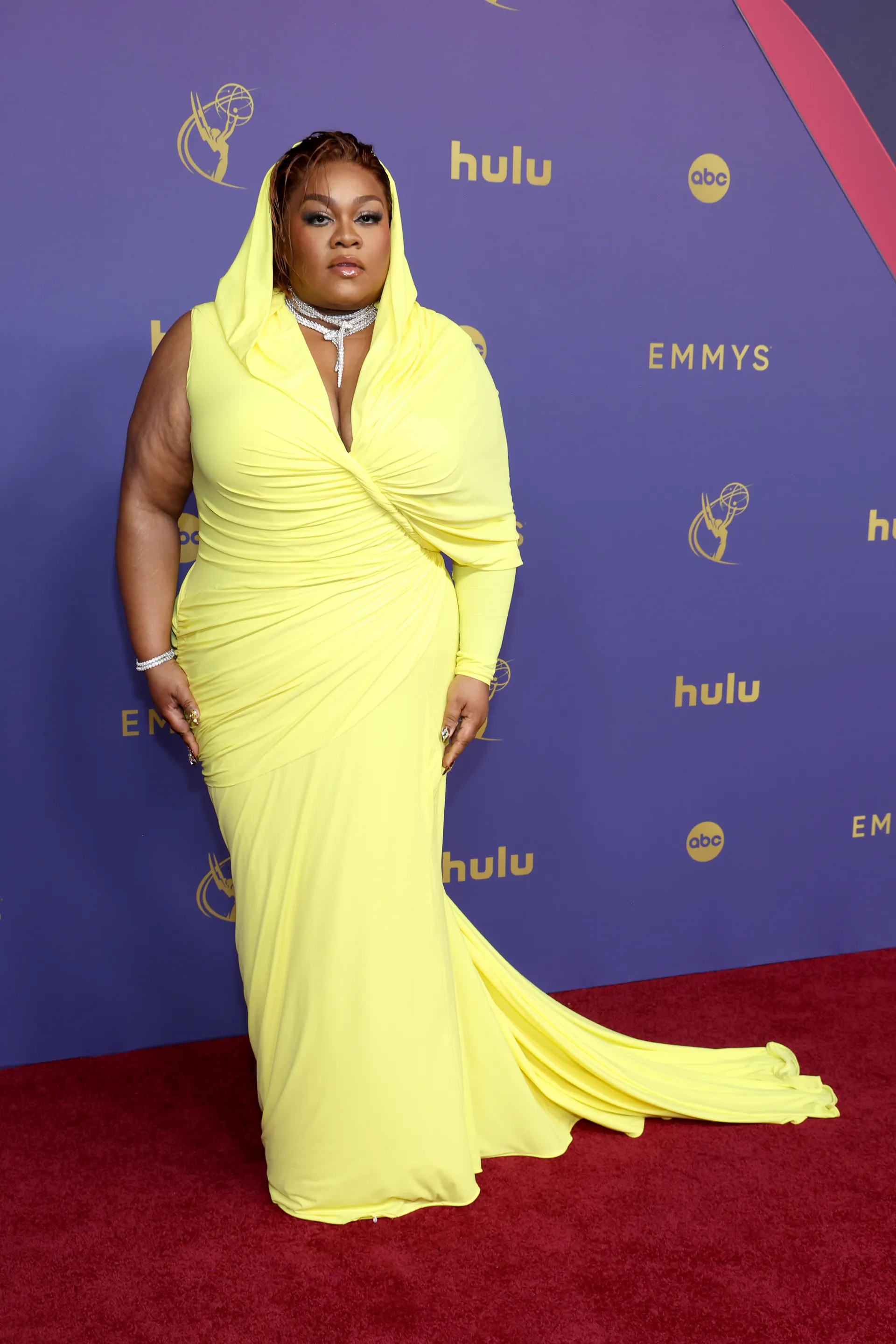 Imagem de mulher em evento de tapete vermelho usando vestido amarelo vibrante com capuz, de tecido drapeado e cauda longa. Emmy 2024.