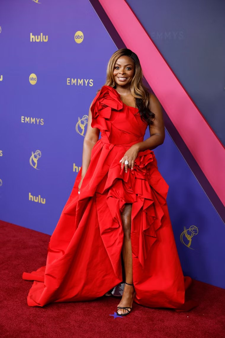 Mulher em evento vestindo um elegante vestido vermelho vibrante, com detalhes volumosos e assimétricos. A peça de um ombro só destaca-se pelo tecido drapeado e camadas elaboradas, seguindo tendências de alta costura. Emmy 2024.