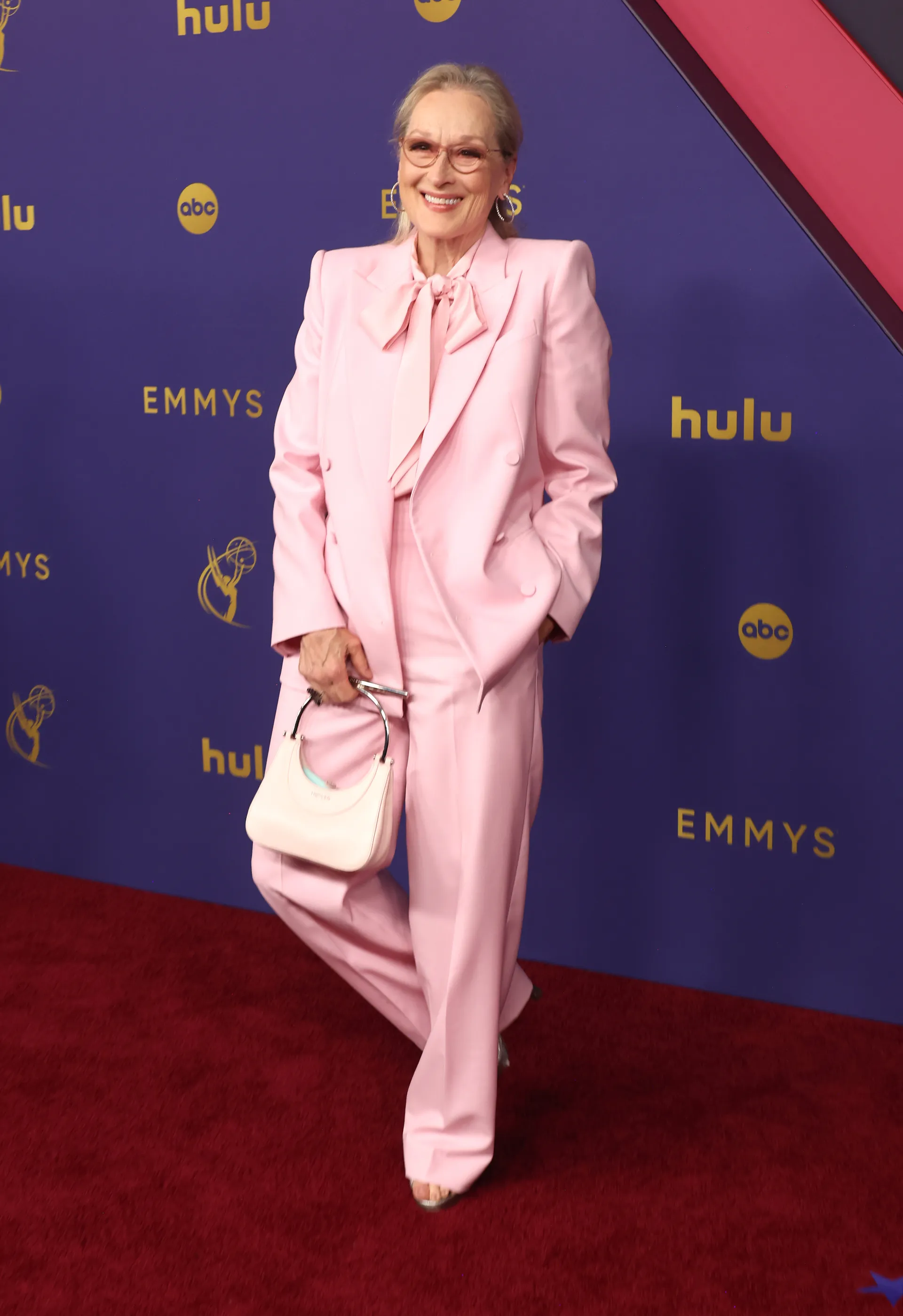 Pessoa usando um elegante terno rosa claro com blazer de ombros marcados e calça de perna larga. O look é complementado por uma camisa com laço no pescoço e uma bolsa de mão pequena, também rosa. Emmy 2024.