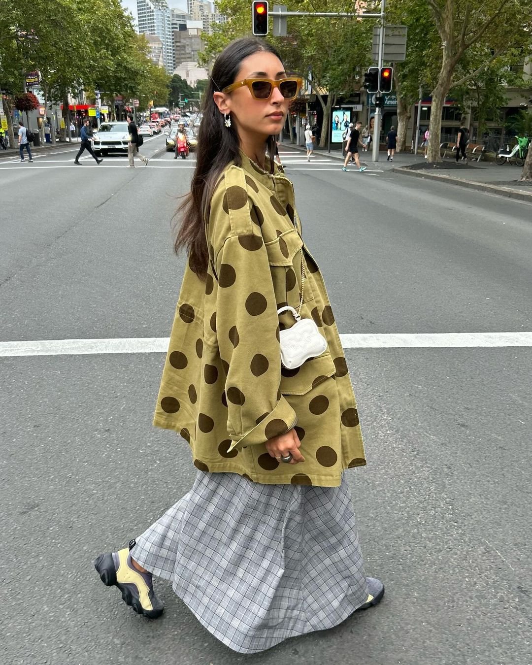 Pessoa caminhando em uma rua da cidade, vestindo uma jaqueta oversized verde oliva com bolinhas grandes, saia longa xadrez cinza e tênis chunky bicolores.