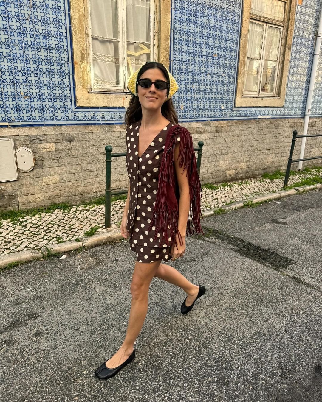 Mulher usando vestido marrom de bolinhas brancas com franjas e sapato preto estilo sapatilha. Complementa o look com acessórios modernos: lenço amarelo na cabeça e óculos de sol.