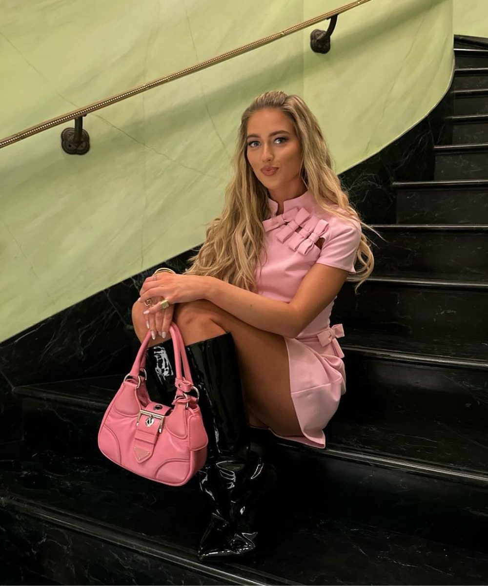 Jovem com vestido rosa curto de gola alta, segurando uma bolsa rosa combinando. Usa botas de couro pretas de cano alto. Look moderno, elegante, enfatizando contrastes de cor e textura. Cabelo solto e maquiagem suave complementam o estilo.