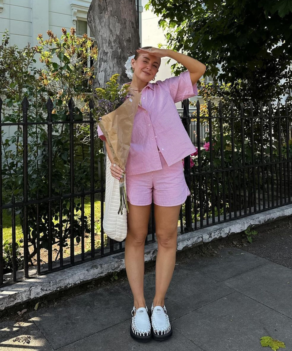 Pessoa usando conjunto de camisa e shorts listrados rosa. Complementa o visual com tênis branco, bolsa de tricô e arranjo de flores. A tendência é casual e confortável, ideal para passeios diurnos. Destaque para a estampa listrada em cores pastel.