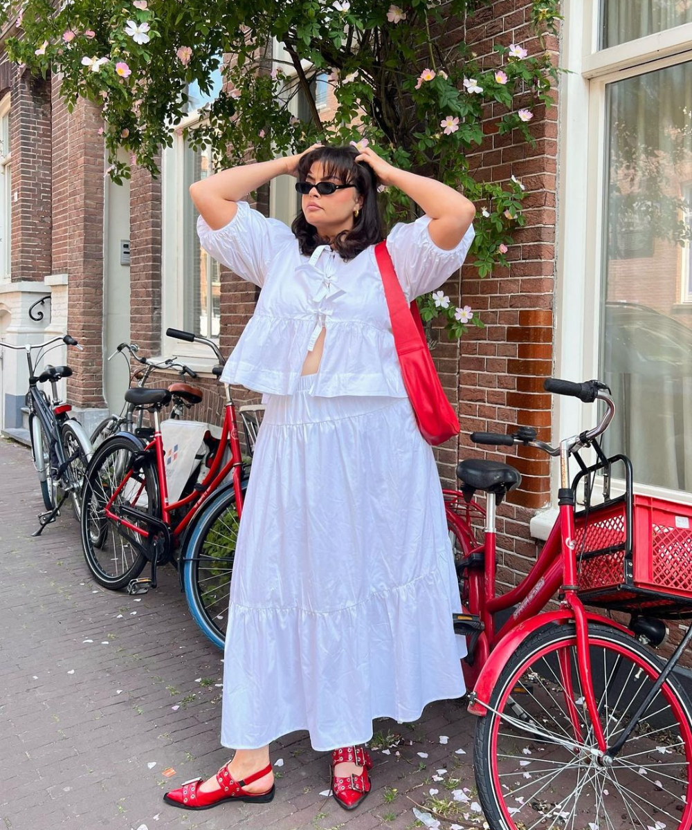 Mulher com vestido branco fluido, estilo boho, usando sandálias vermelhas combinando com uma bolsa vermelha de ombro. Óculos de sol pequenos e escuros completam o look, destacando uma tendência casual e confortável com toque de cor.