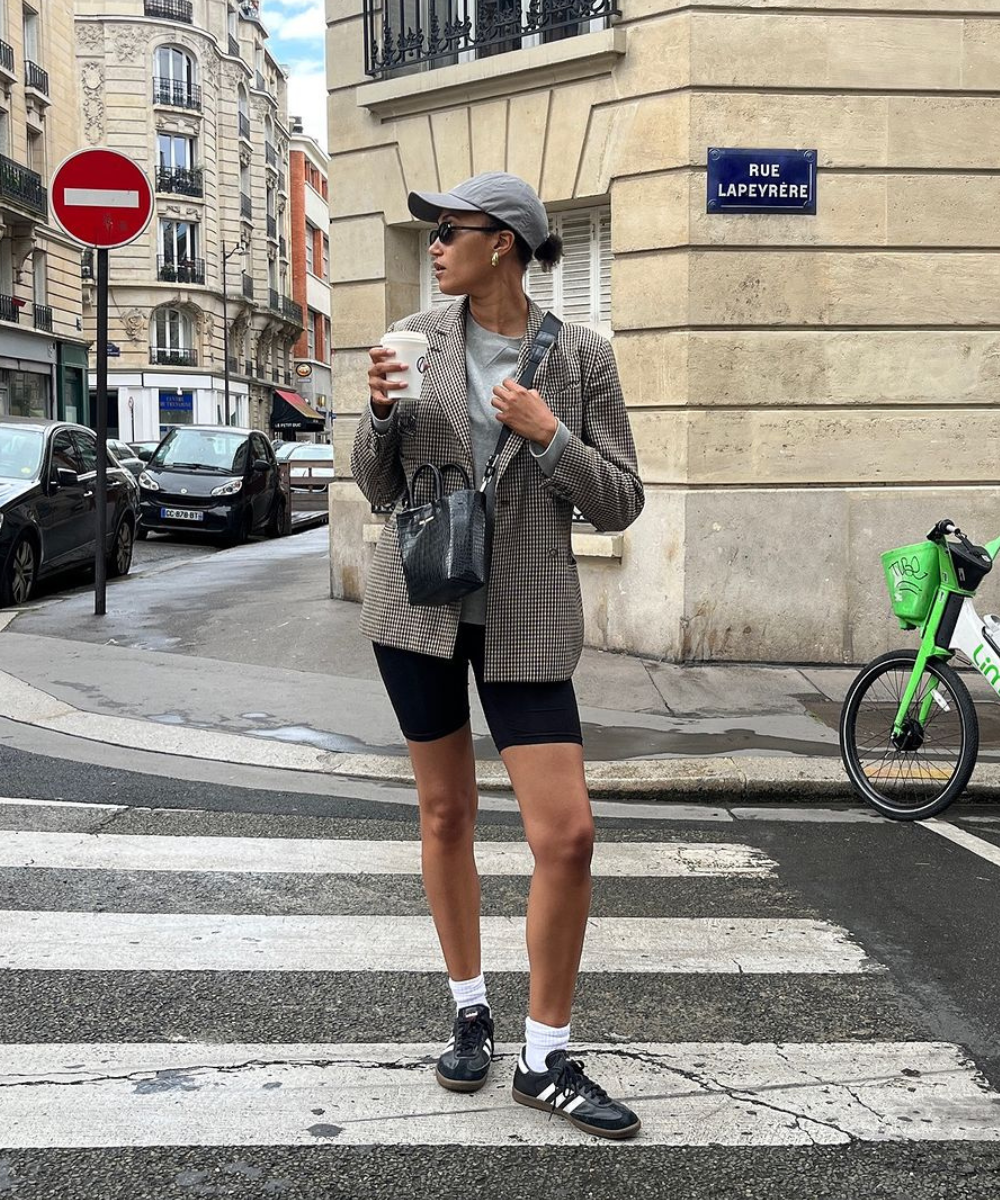 Pessoa usando blazer xadrez com bermuda ciclista preta, camiseta neutra, tênis preto, meias brancas e boné cinza. Acessórios incluem bolsa preta e óculos de sol. Estilo urbano casual com toque esportivo. Foto em uma rua parisiense.