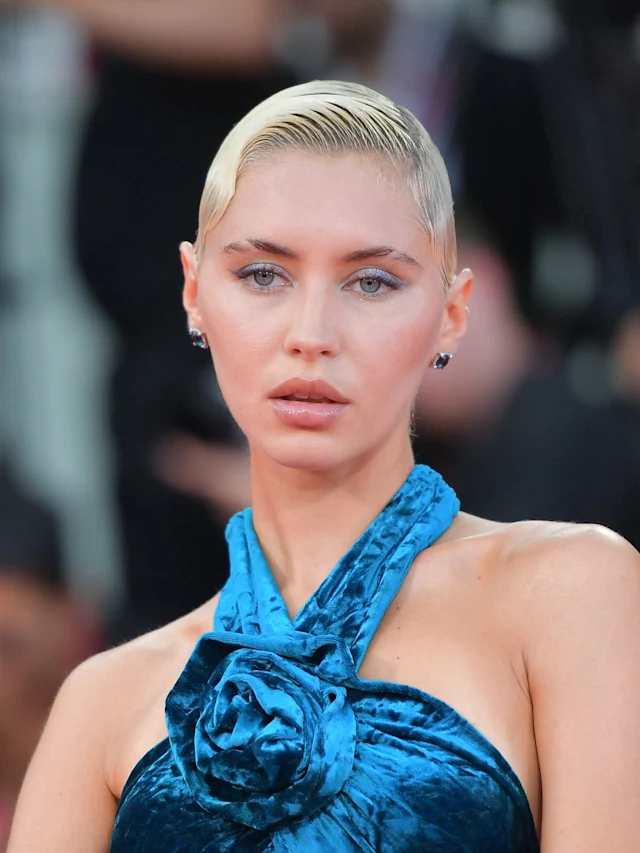 Mulher com cabelo loiro platinado, penteado liso e colado, usando vestido de veludo azul com detalhe em flor no pescoço. Se destaca pelo estilo sofisticado e moderno, combinando elegância clássica com tendências  contemporâneas. festival de Veneza