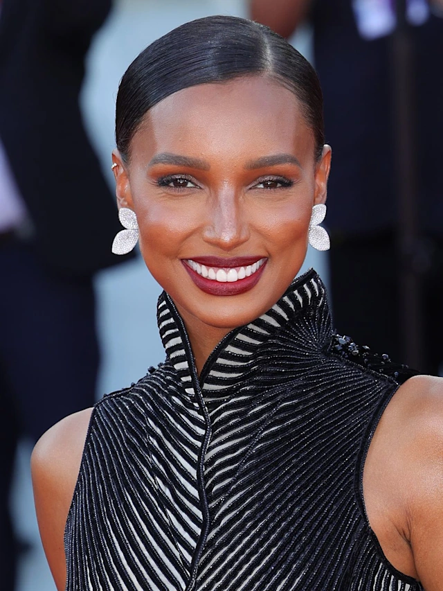 Mulher com look elegante, usando vestido preto com detalhes texturizados e gola alta. O cabelo está preso em um coque liso e elegante, e ela usa brincos grandes e brancos. Maquiagem sofisticada com batom vermelho. festival de veneza