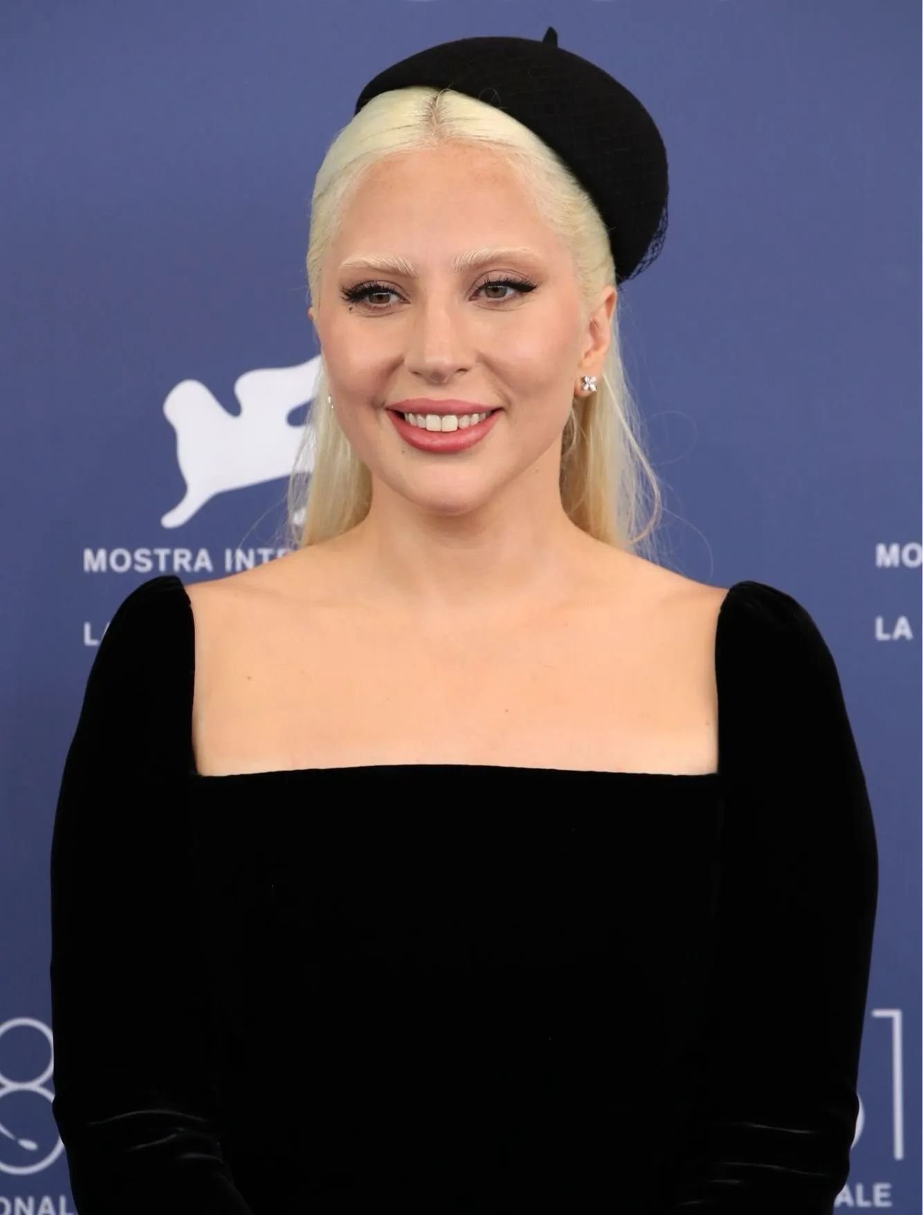 Mulher usando vestido preto de veludo com decote quadrado e manga longa. Ela complementa o look com um chapéu preto estiloso e brincos de diamante. festival de Veneza