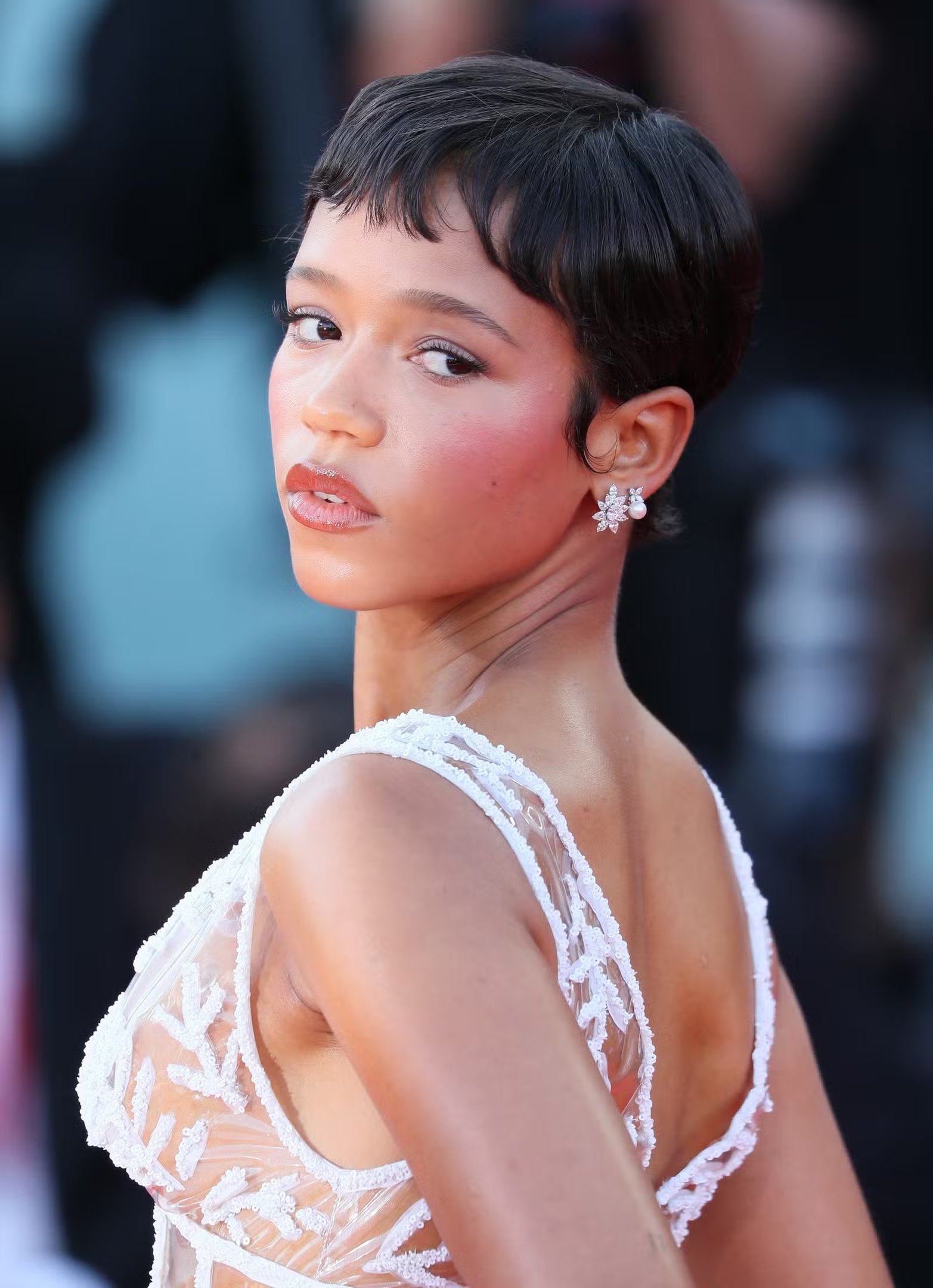 Imagem de uma mulher com cabelo curto e liso, usando um vestido branco de alças finas, bordado com detalhes florais transparentes, e brinco delicado. festival de Veneza