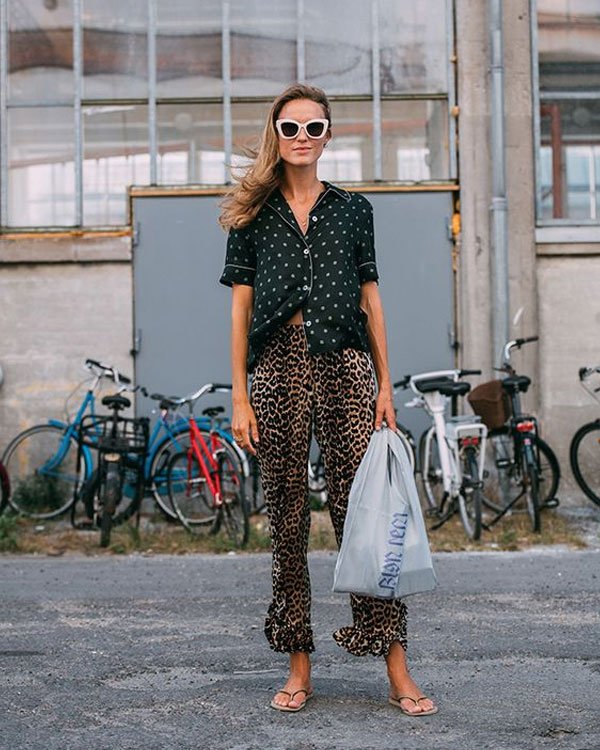 Mulher estilosa usa óculos grandes brancos, blusa preta com bolinhas brancas e calça estampa animal print. Completa o look com sandálias rasteiras e bolsa plástica, misturando tendências de moda retrô e moderna.