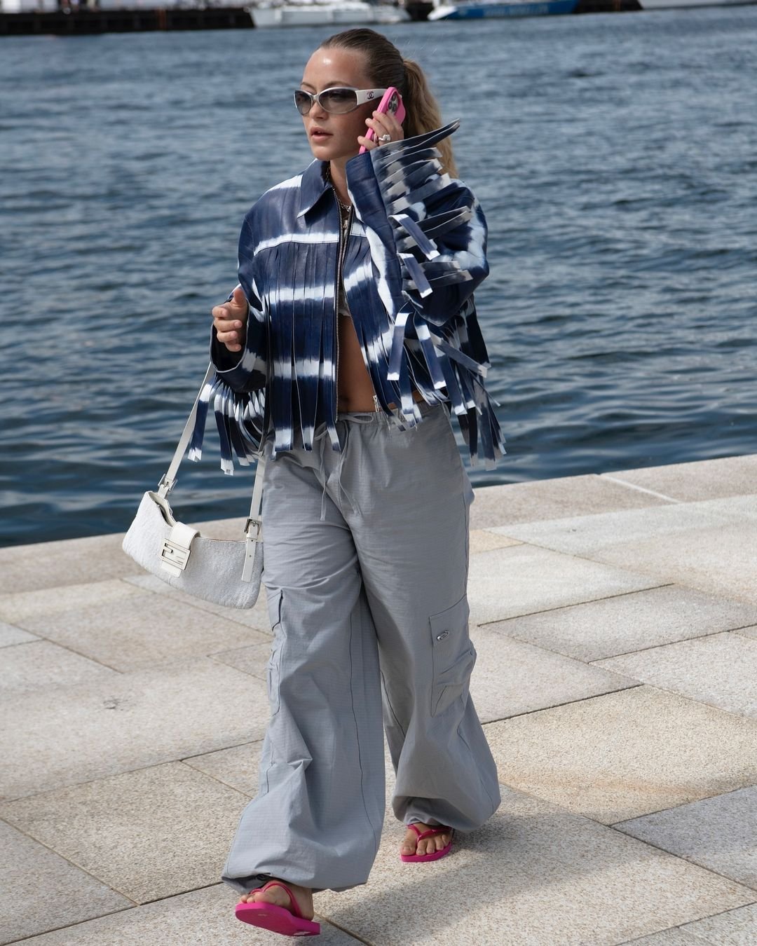 Mulher usando jaqueta de franjas azul e branca com calça cargo cinza, sandálias rosa e carregando uma bolsa branca. Óculos de sol modernos e celular rosa complementam o visual,.