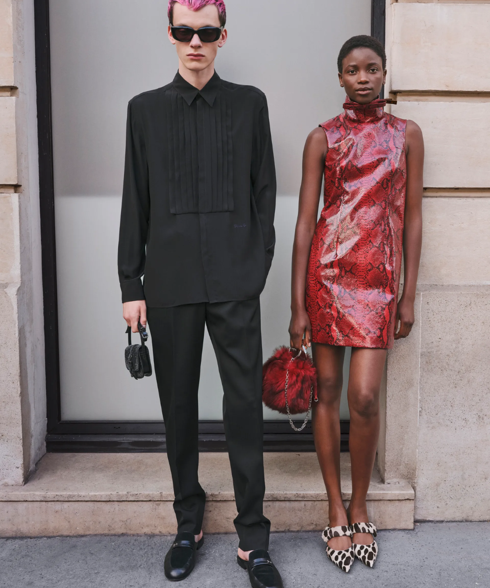 Dois modelos posando em frente a um prédio. O homem veste camisa social preta e calça preta, completando o look com sapatos pretos e cabelos rosa. A mulher usa um vestido curto de cor vermelha com estampa de cobra, sapatos de salto com estampa animal e bolsa de pelúcia vermelha.