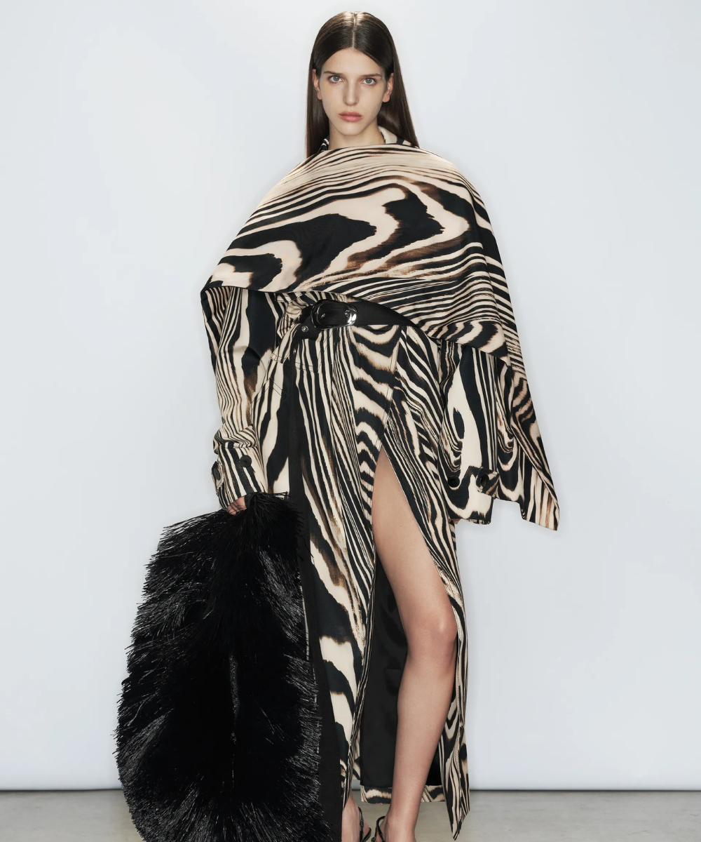 Imagem de uma mulher vestindo um vestido longo com estampa animal print em preto e bege, acompanhado de um cinto preto marcando a cintura. A fenda alta e um enorme acessório preto completam o look contemporâneo e ousado, destacando tendências modernas e elegantes.