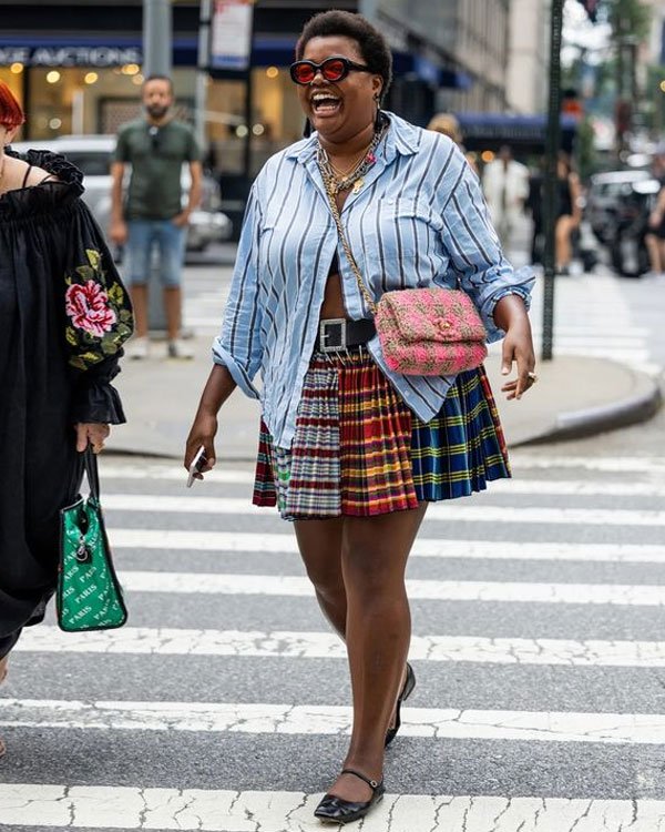 Mulher no centro da cidade vestindo uma camisa listrada azul, saia plissada de estampa xadrez multicolorida e sapatos pretos.