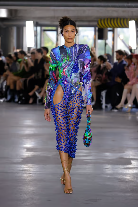 Modelo vestindo look futurista e ousado, composto por vestido de malha azul brilhante com cortes assimétricos e detalhes texturizados, combinado com top de manga longa estampado abstrato multicolorido. Semanas de moda internacionais.