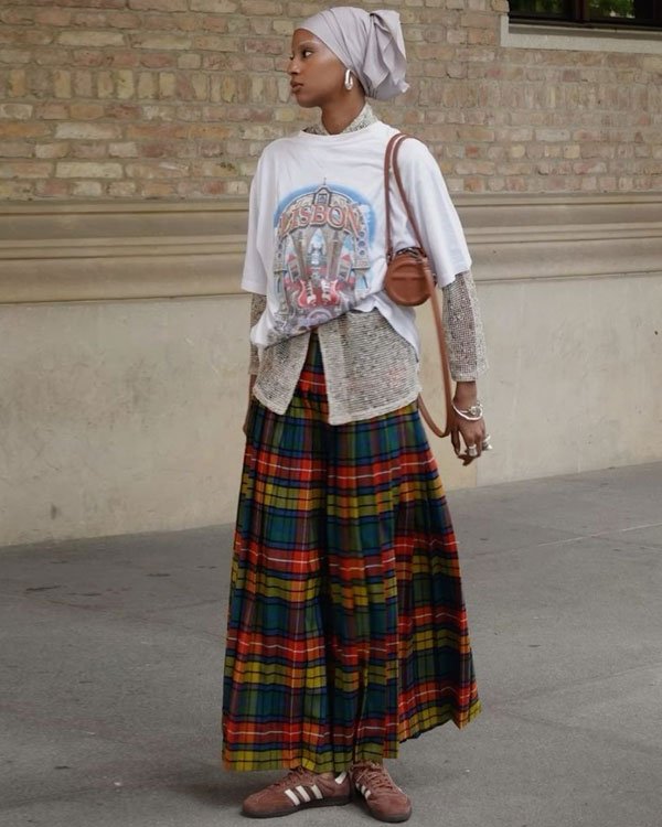 Pessoa com camiseta oversized gráfica, blusa de tricô por baixo, saia longa com estampa xadrez vibrante, tênis vintage Adidas e bolsa de couro marrom. Acessórios incluem brincos grandes e pulseiras.