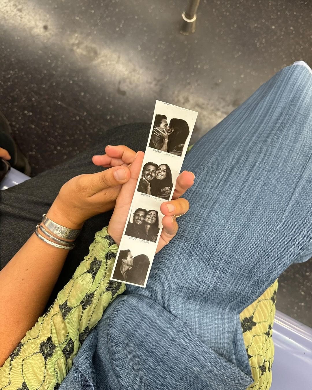 Foto de um casal segurando tiras de fotos em um metrô. Ela veste blusa verde com textura diferenciada e mangas bufantes, ele com calça escura. Estilos modernos e casuais, tendências de moda urbana.