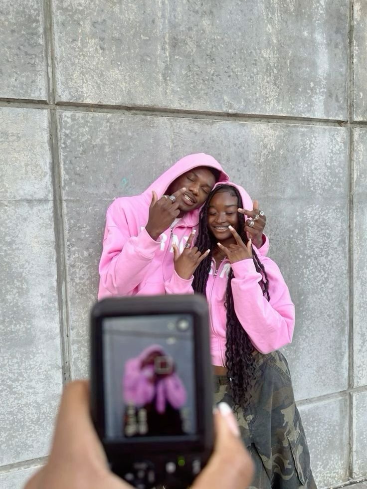 Casal vestindo moletons rosa combinando, destacando uma tendência casual e confortável de moda. Eles posam em frente a uma parede de concreto, com um blend de estilos urbanos e streetwear. A foto é capturada através de uma câmera digital.