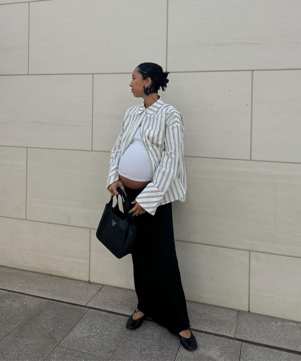 Grávida com camisa listrada oversized e calça preta folgada. Bolsa preta de couro e sapatos pretos confortáveis. Estilo casual chique, combinando tendências de conforto com elegância. Cabelo preso e acessórios discretos completam o look.