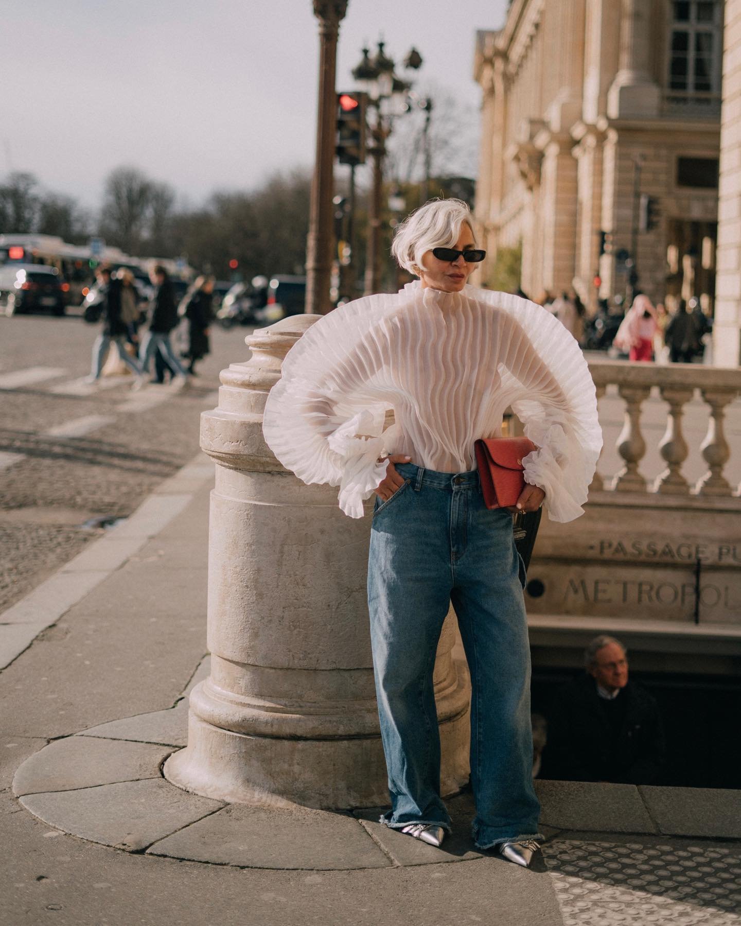 Grece Ghanem usa blusa branca semitransparente com mangas volumosas de tecido plissado, combinada com jeans de modelagem reta e sapatilhas prateadas. Óculos escuros e carteira vermelha completam o visual.