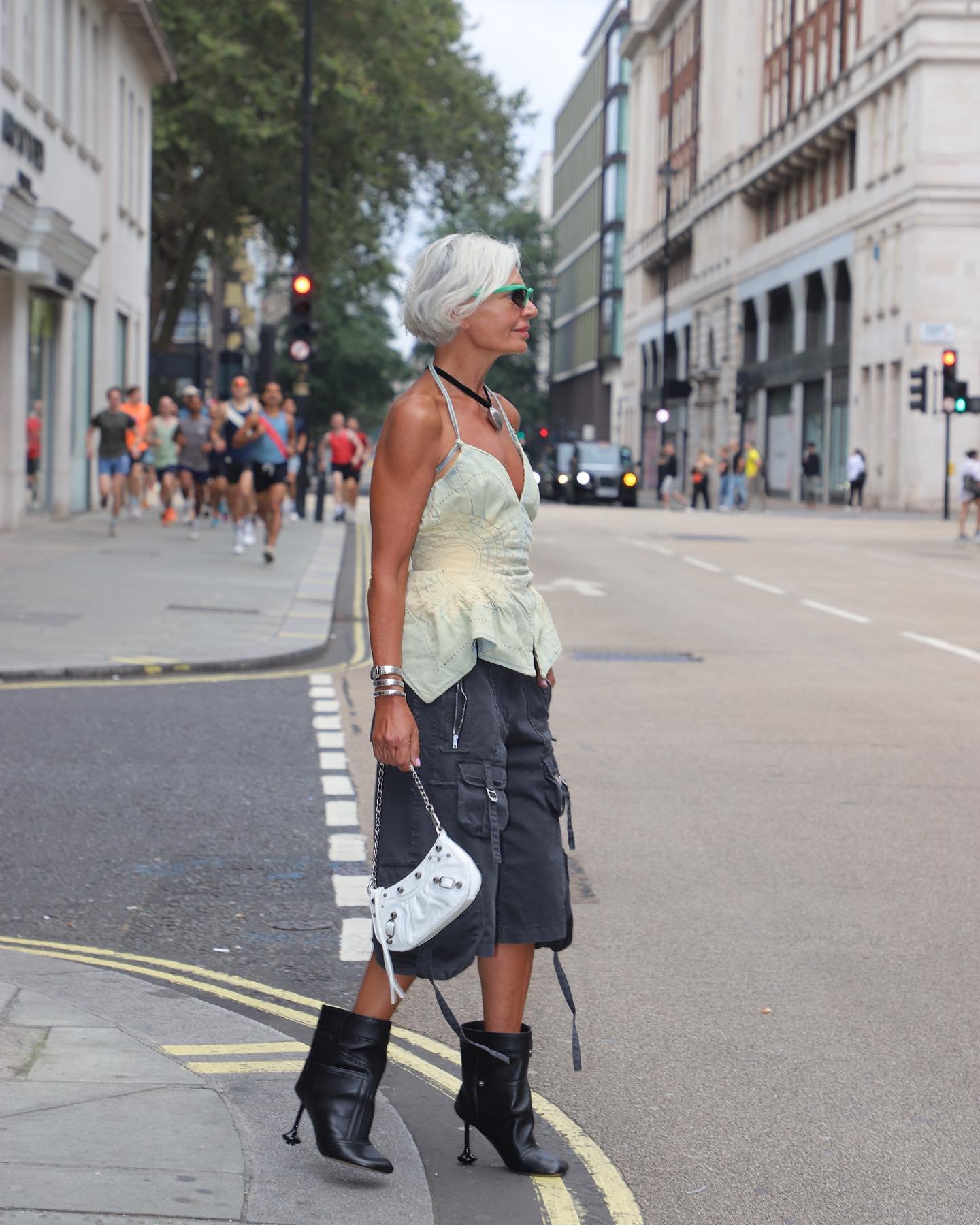 Mulher de cabelo grisalho e corte bob, usando top assimétrico verde claro, bermuda cargo preta, botas de cano médio pretas e bolsa branca com corrente. Óculos de sol modernos e pulseiras completam o look. Estilo urbano e contemporâneo.