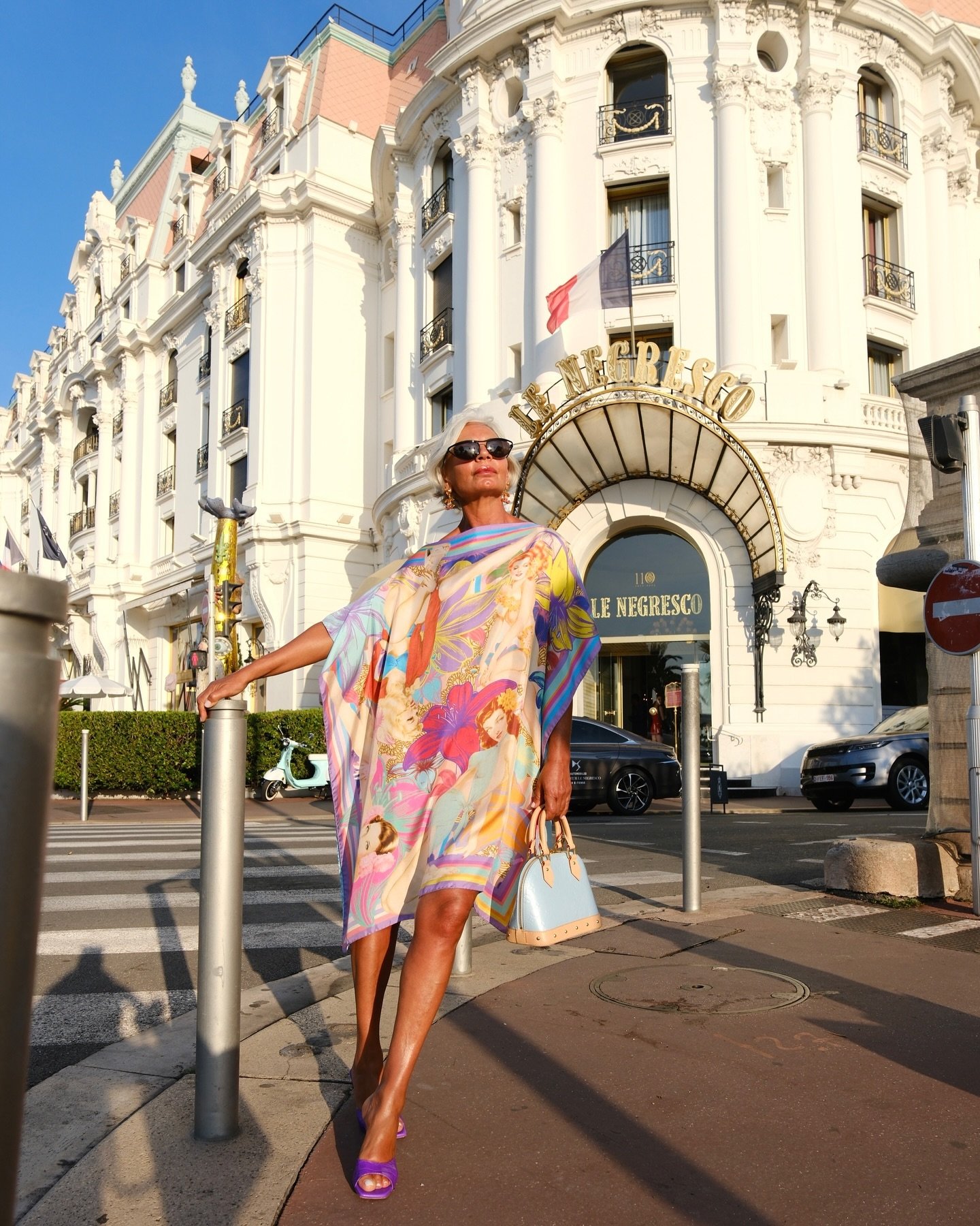 Mulher de cabelos brancos, usa vestido largo e estampado com cores vivas e imagens de seres mitológicos. Acessórios incluem óculos escuros, sandálias roxas e bolsa estruturada em tom pastel. Cenário de fundo é um hotel clássico.