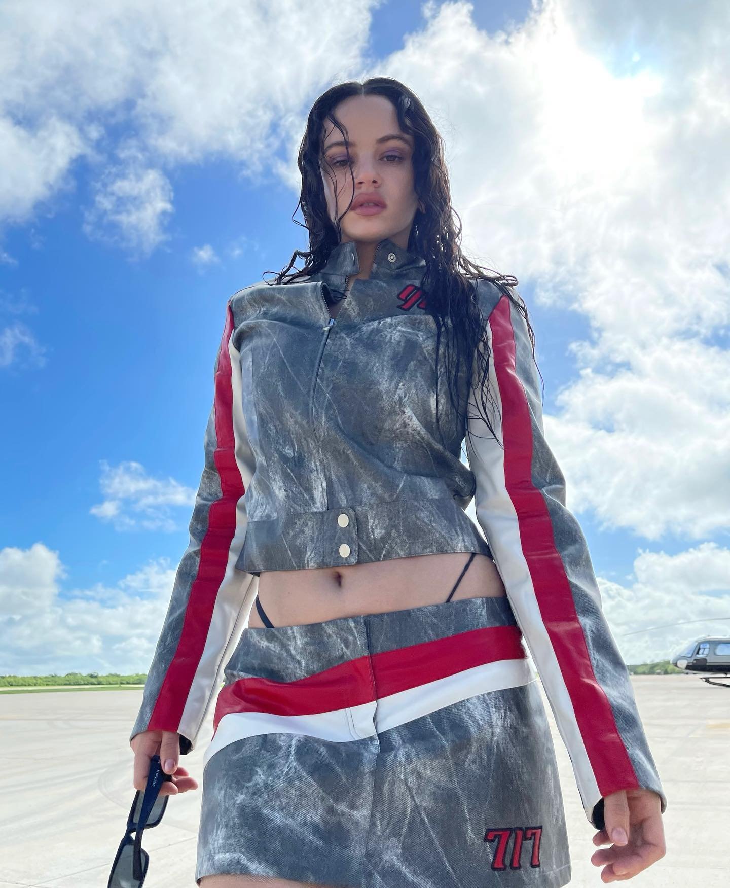 Mulher vestindo conjunto cropped e minissaia de design moderno com padrão marmorizado cinza, listras vermelhas e brancas, e detalhes assimétricos. O look é complementado por óculos de sol estilo oversized. Tendência de moda urbana e futurista.