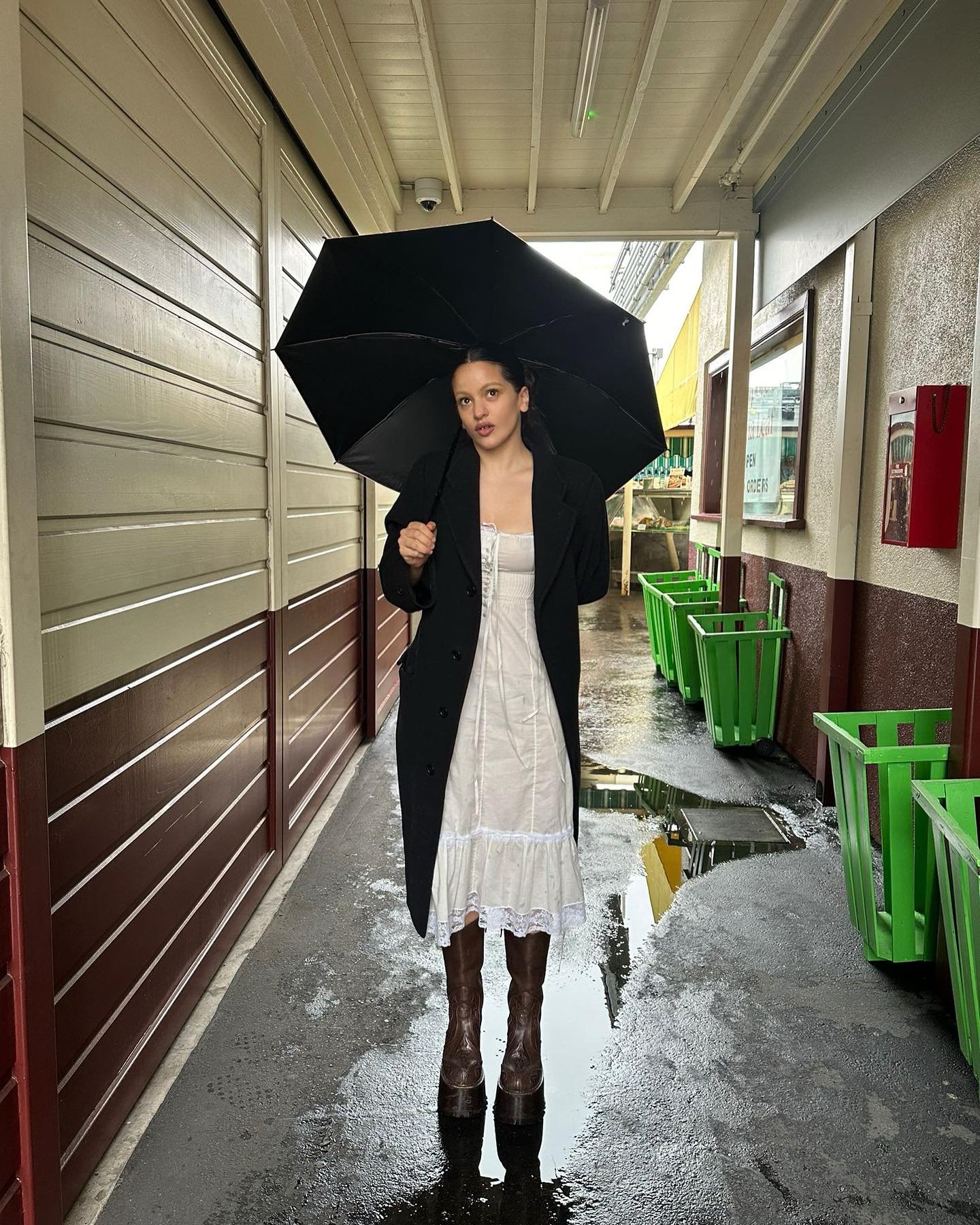 Pessoa usando vestido branco estilo vintage, casaco preto oversized e botas marrons de cano alto. Segura um guarda-chuva preto em uma passagem coberta com chão molhado. Combinação de elementos clássicos e contemporâneos, destacando tendêncais de moda outono/inverno.