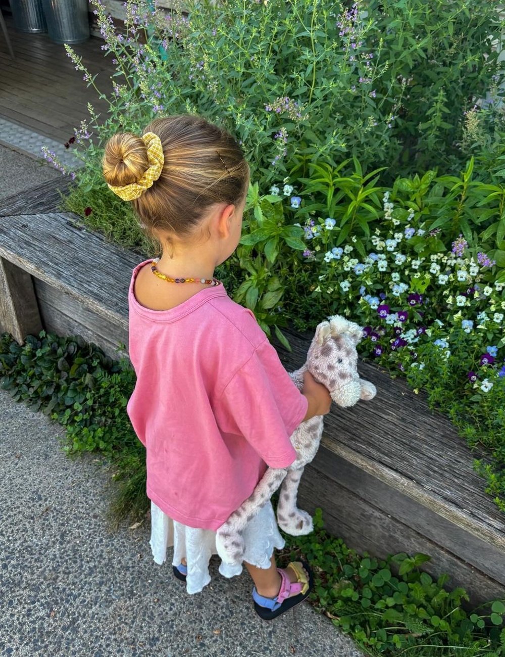 Criança de costas, usando camiseta rosa ampla, saia branca rendada e sandálias coloridas. Cabelo preso em coque com scrunchie amarelo. Segura um brinquedo de pelúcia cinza. Influencer de moda infantil.