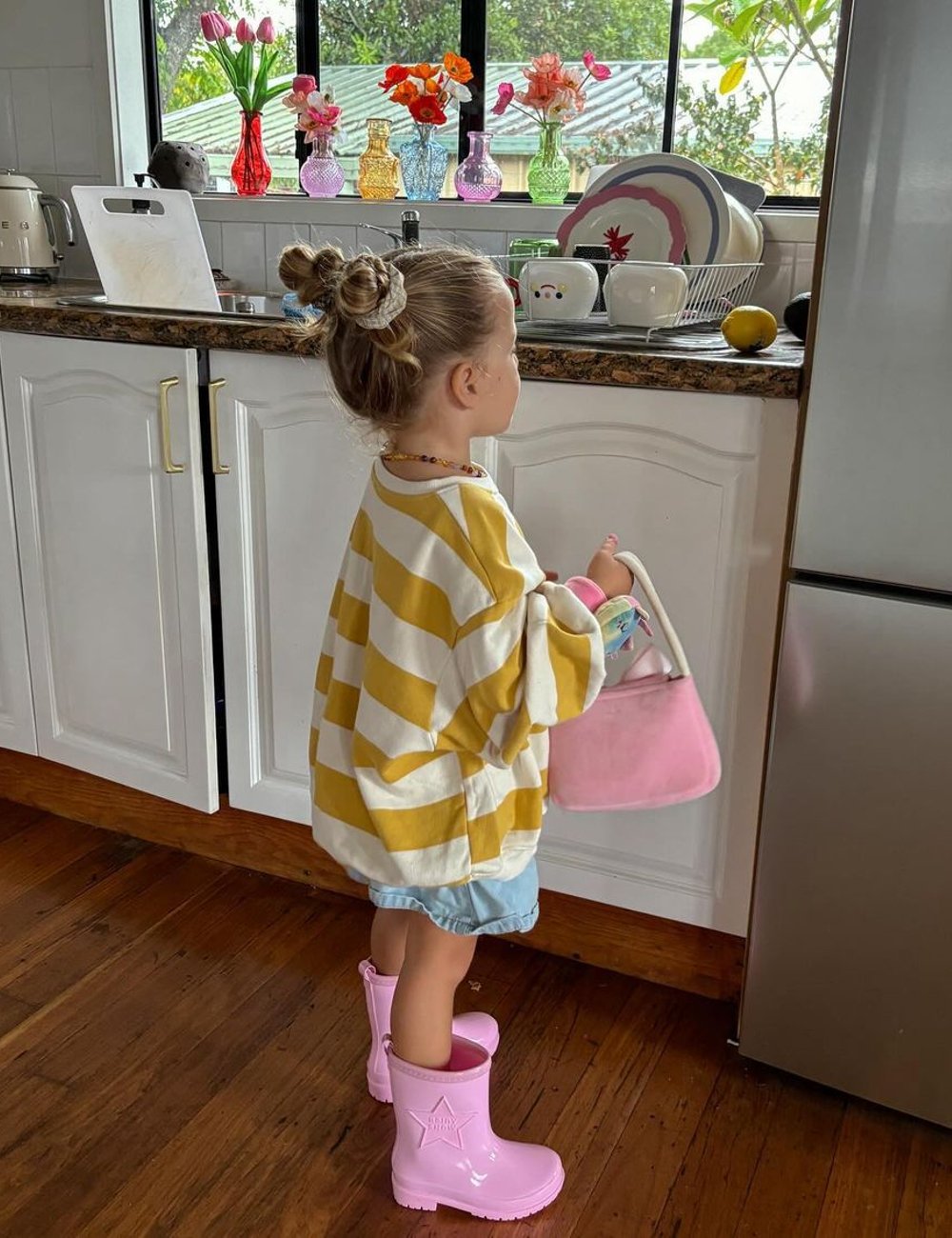 Criança veste camiseta listrada oversized amarela e branca, short jeans e botas de chuva rosa. Estilo descontraído e casual, destacando-se pelo uso de tendências infantis modernas como acessórios de cabelo estilizados. Influencer de moda infantil.