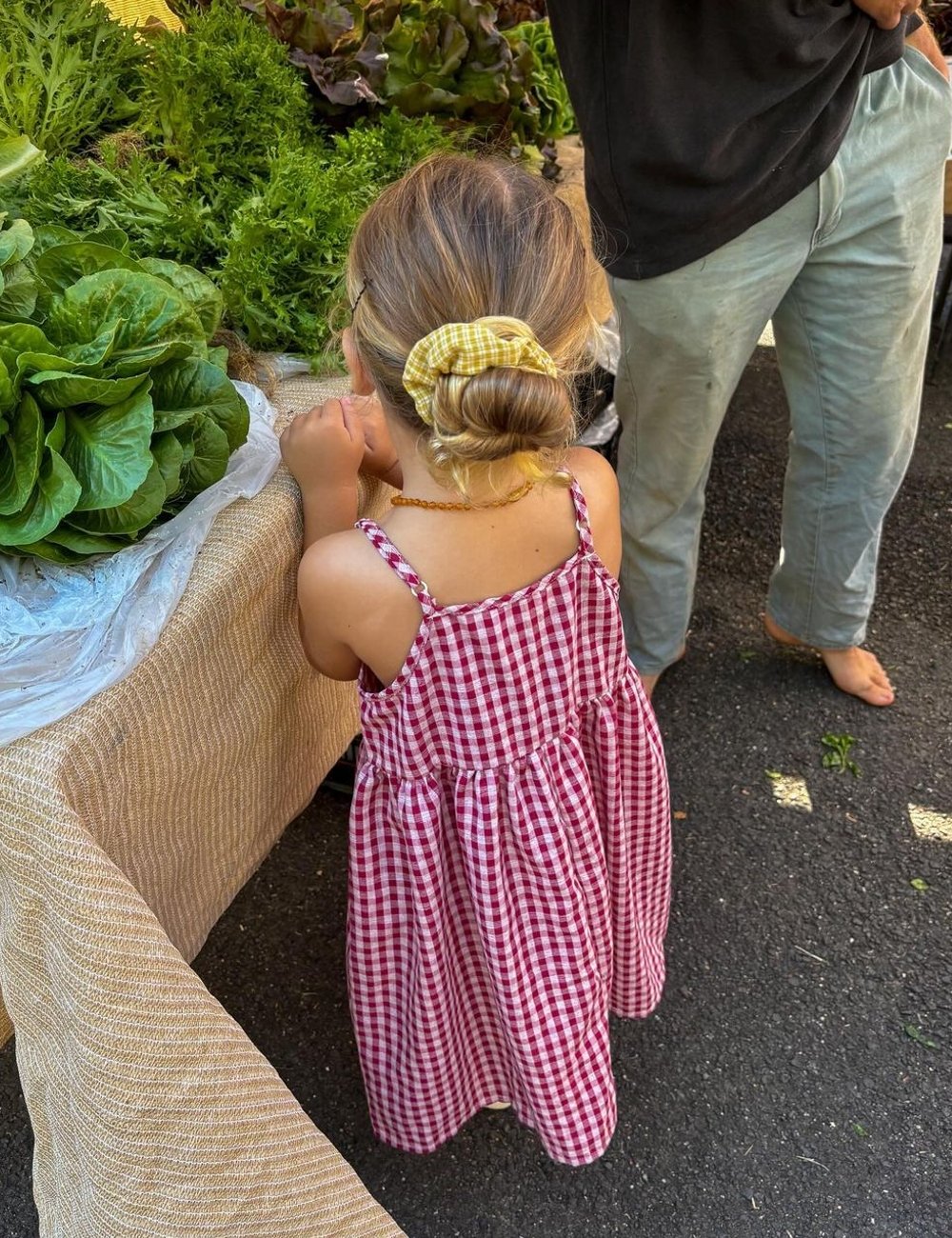 Criança com vestido xadrez vermelho e branco estilo vintage, cabelo preso com laço amarelo, em feira ao ar livre. Roupa casual e fofa, perfeita para dias ensolarados e passeios informais, destacando influencer de moda infantil.