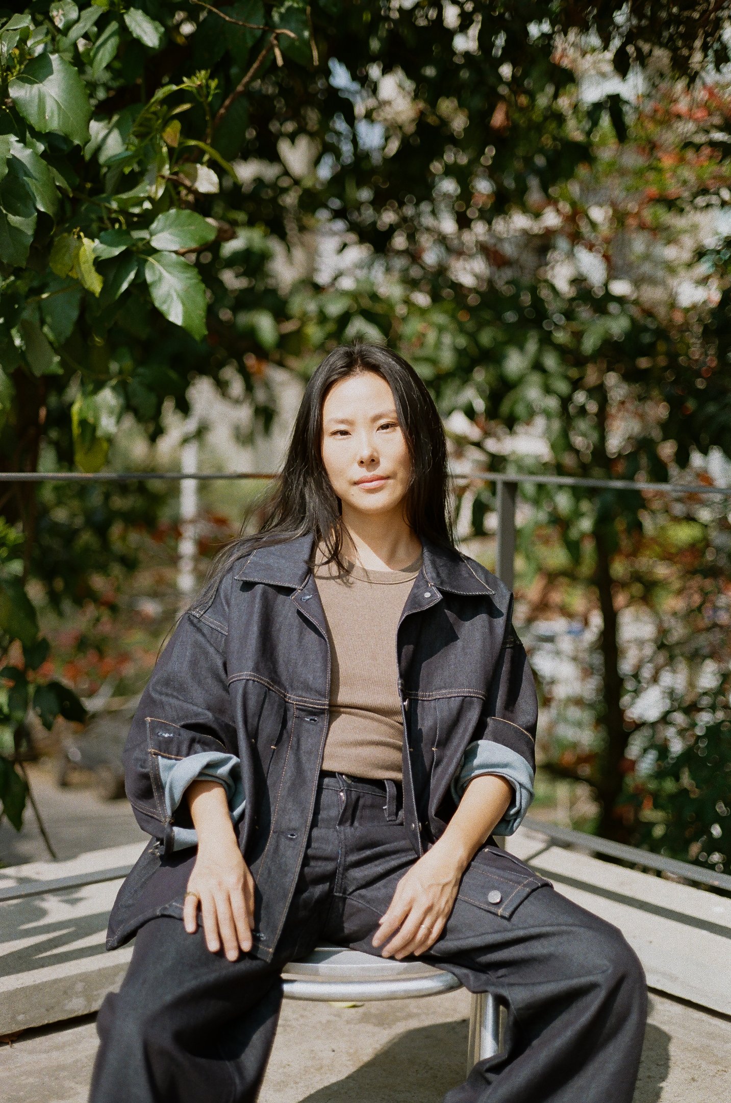 Pessoa com estilo casual e contemporâneo. Ela veste uma jaqueta oversized jeans combinada com uma camiseta marrom e calça jeans de cintura alta. As mangas da jaqueta estão dobradas, conferindo um ar descontraído e moderno.