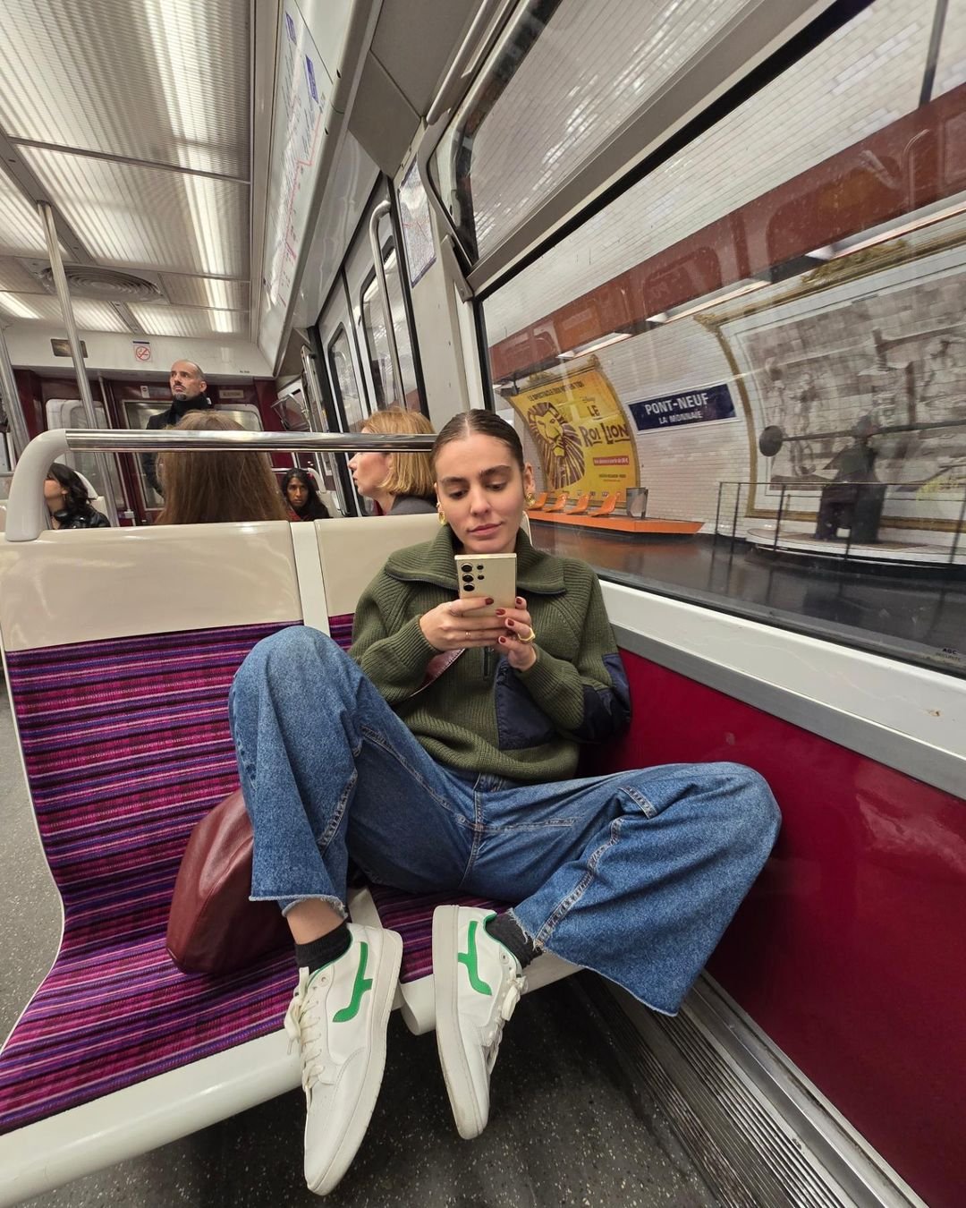 Mulher ecenta no metrô veste suéter verde oversized, calça jeans larga, e tênis branco com detalhe verde. Estilo casual e confortável, combinando tendências de moda urbana e relaxada. Cenário de transporte público europeu.