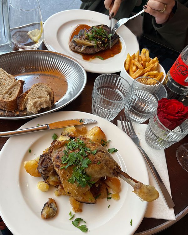 Almoço em restaurante sofisticado. Pratos gourmet com apresentação cuidadosa incluem carnes, batatas e pães; bebida e coca-cola acompanham. Tendência de moda: jaqueta militar verde e unhas escuras, acessórios minimalistas.