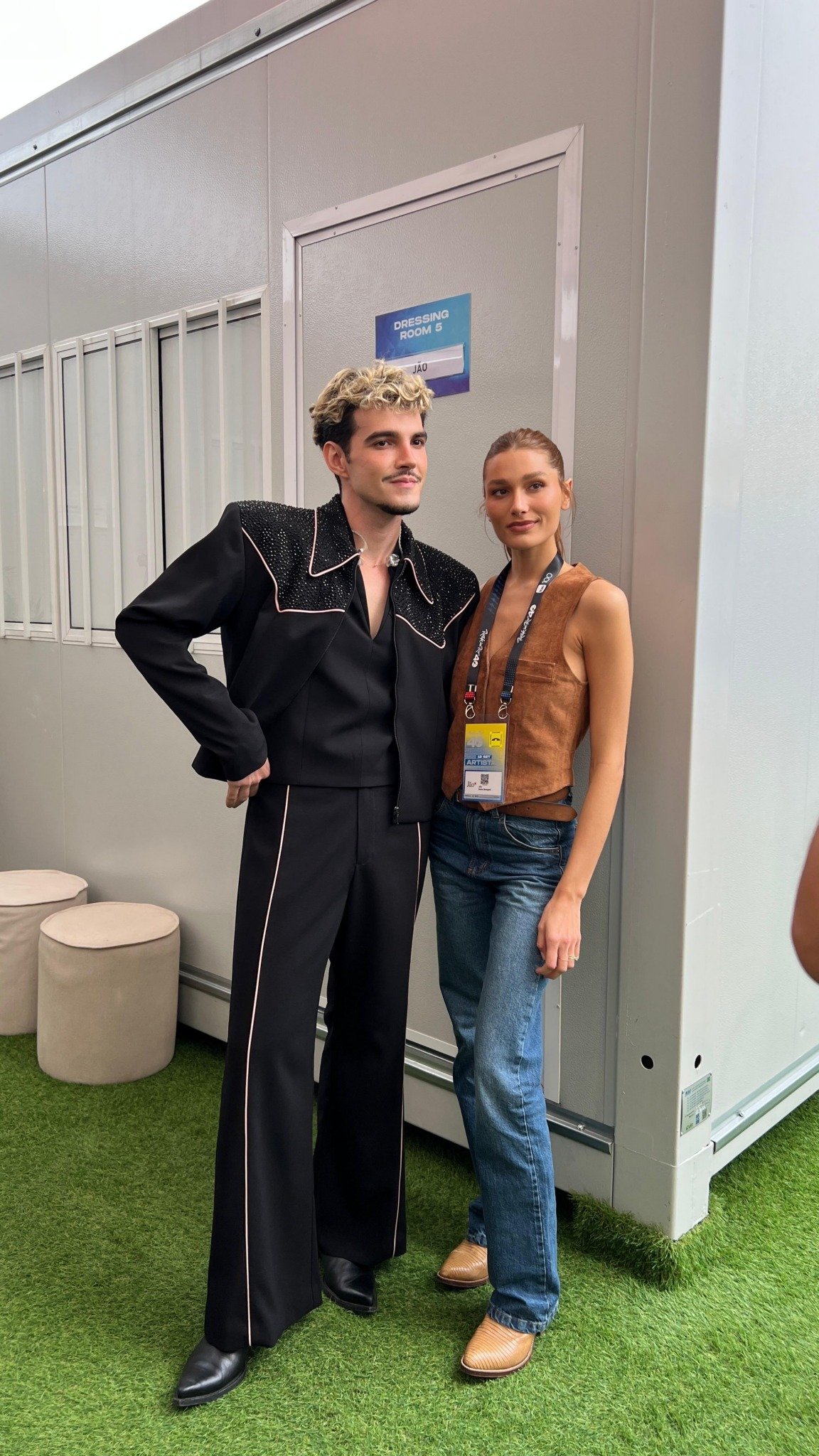 Jão e Sasha em frente a um camarim do Rock in Rio. Uma veste um conjunto preto com detalhes claros e ombros adornados, tendência glam. A outra usa colete de camurça marrom, jeans e botas, estilo casual e rústico.