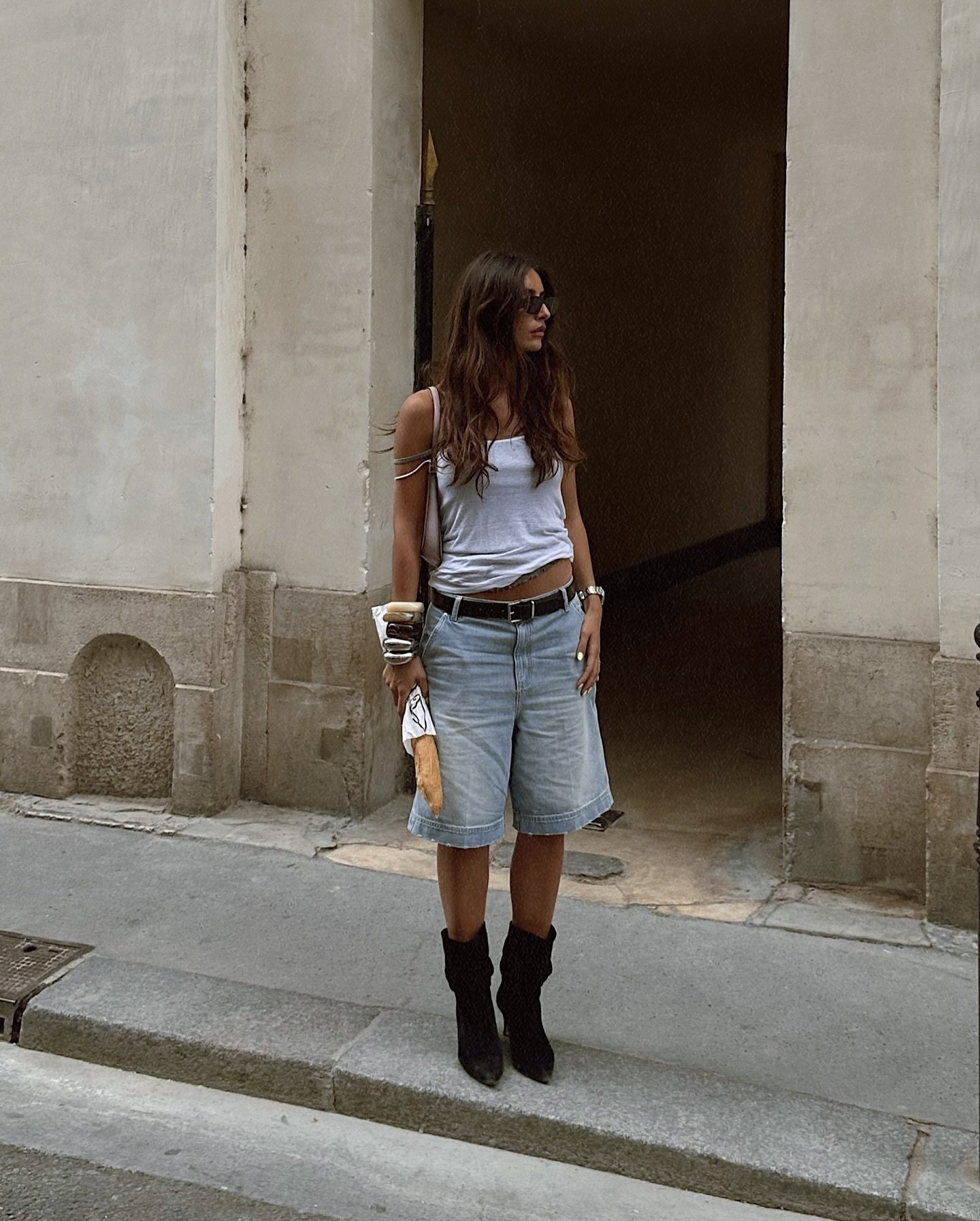 Mulher com estilo casual de verão, vestindo bermuda jeans, regata branca e botas pretas. Óculos de sol escuros e acessórios boho completam o look, como pulseiras e uma bolsa pequena, além de um pão francês nas mãos.