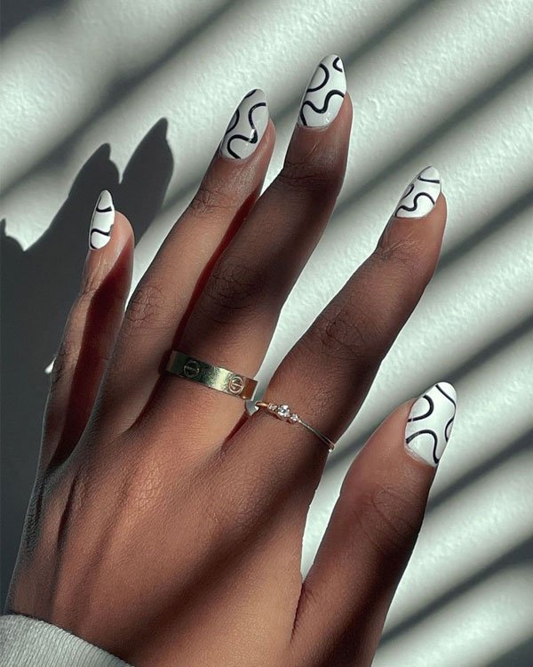 Mão com unhas longas e decoradas com esmalte branco e linhas pretas abstratas. Uso de anéis delicados, um dourado grosso e outro prateado fino, criando um visual elegante e moderno.