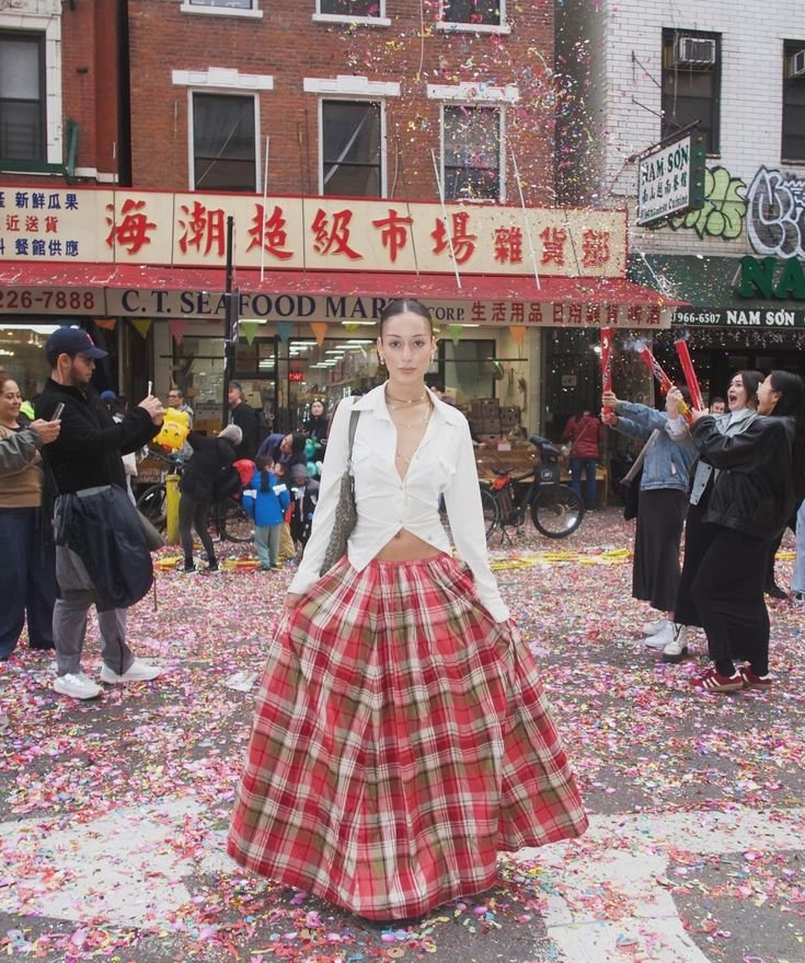 Mulher usando saia xadrez longa em vermelho e branco, combinada com blusa branca aberta e ajustada. Estilo atual mescla elementos tradicionais com toque moderno. Fundo de celebração urbana com confetes.