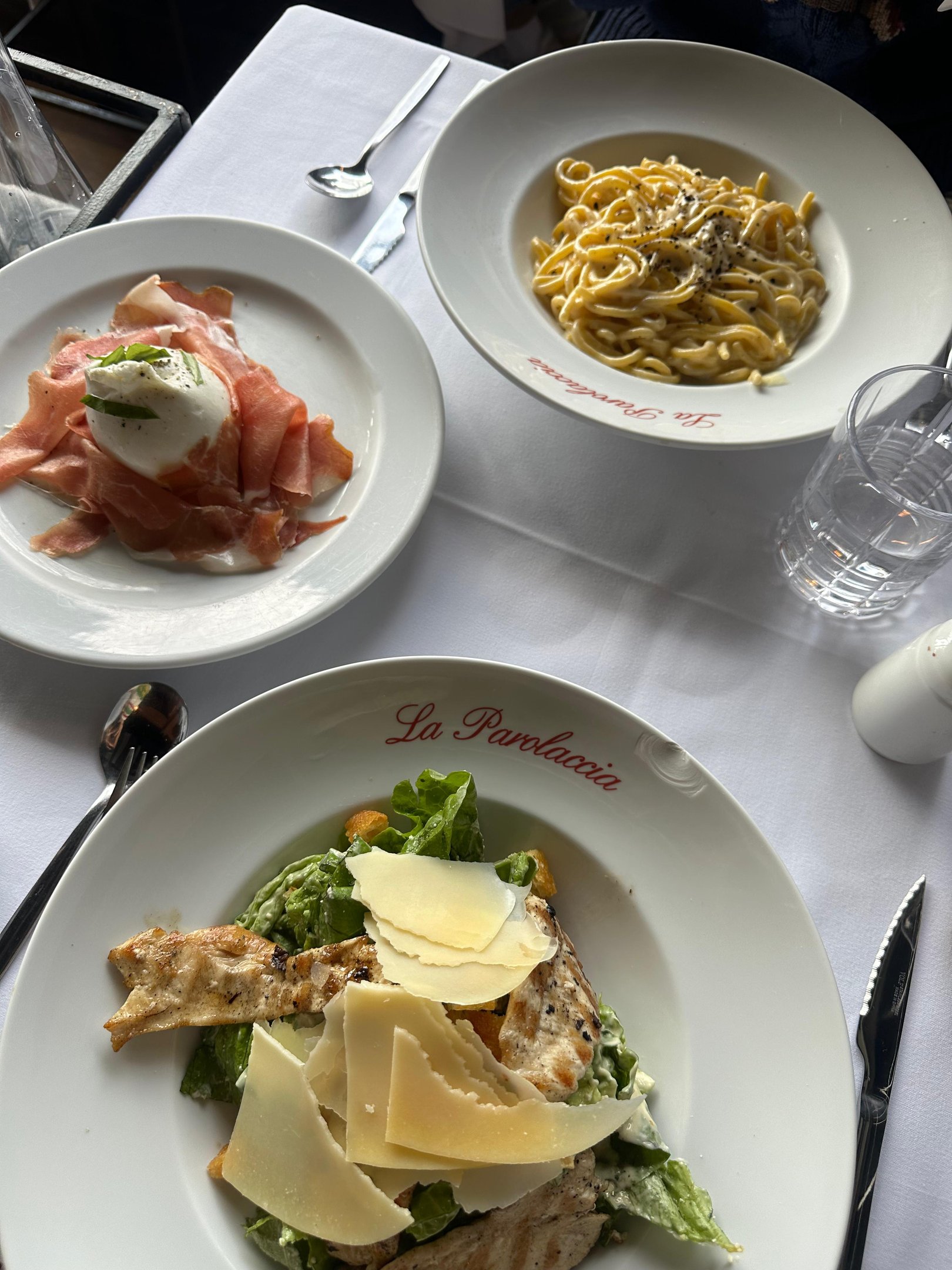 Refeição refinada no restaurante La Parolaccia, com pratos de presunto e queijo burrata, salada Caesar com lascas de parmesão e prato de massa com molho branco. Mesa elegante com toalhas brancas e louça sofisticada. Restaurantes em Buenos Aires.