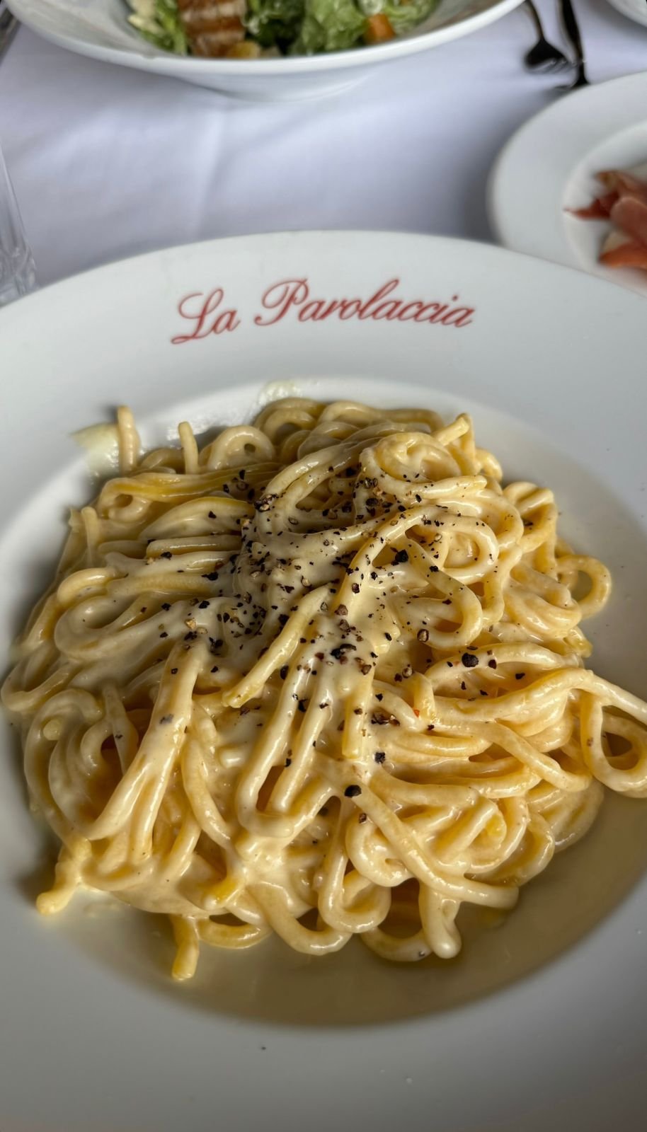 Prato de espaguete ao molho branco com pimenta-do-reino, servido em um restaurante italiano elegante. Ao fundo, uma salada verde e fatias de presunto cru em pratos brancos. Ambiente refinado, ideal para amantes da culinária italiana.