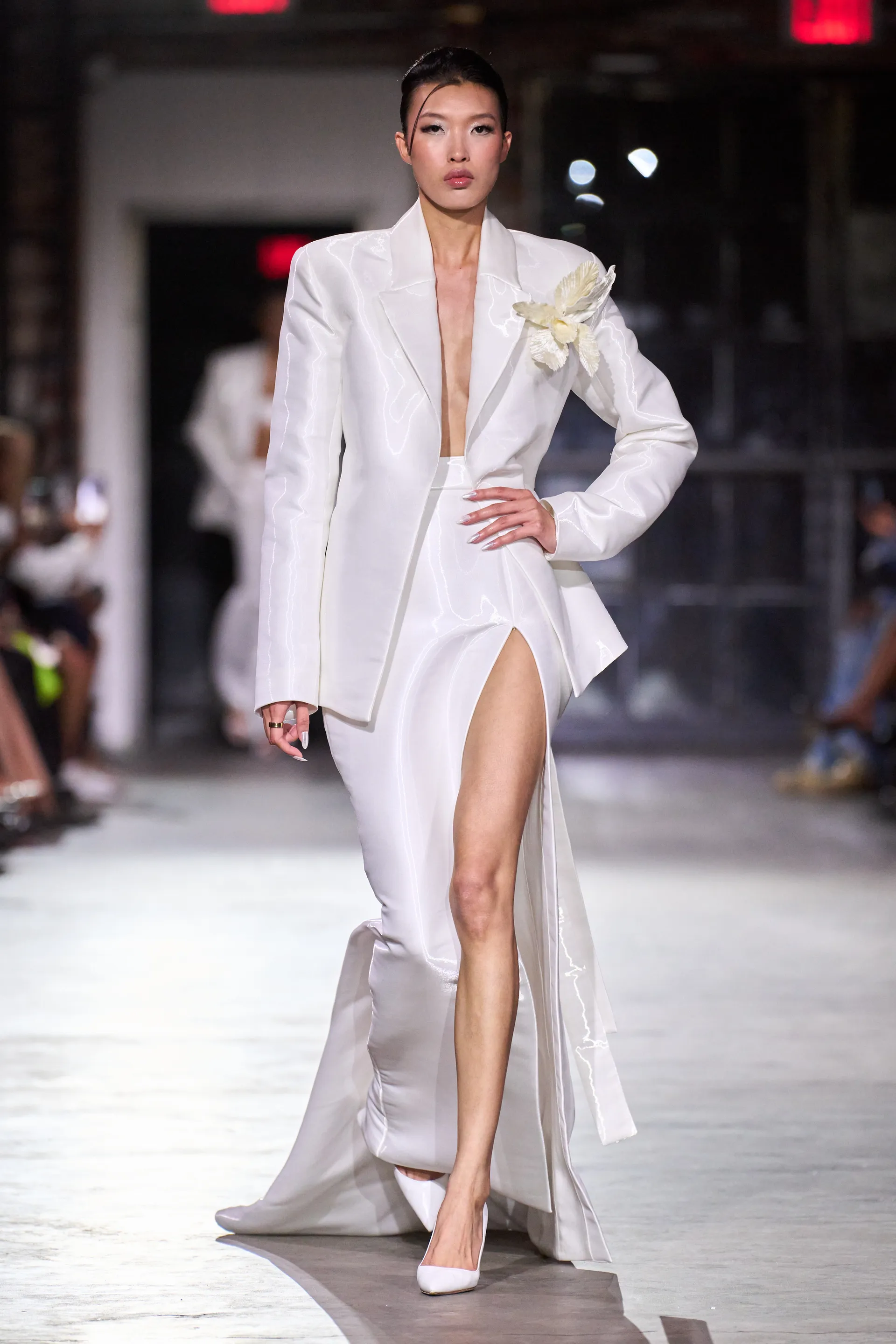 Modelo usando um look elegante de alta-costura com blazer oversized branco e saia longa com fenda alta. O visual monocromático é complementado por um broche floral no ombro e sapatos de salto alto. NYFW SS25