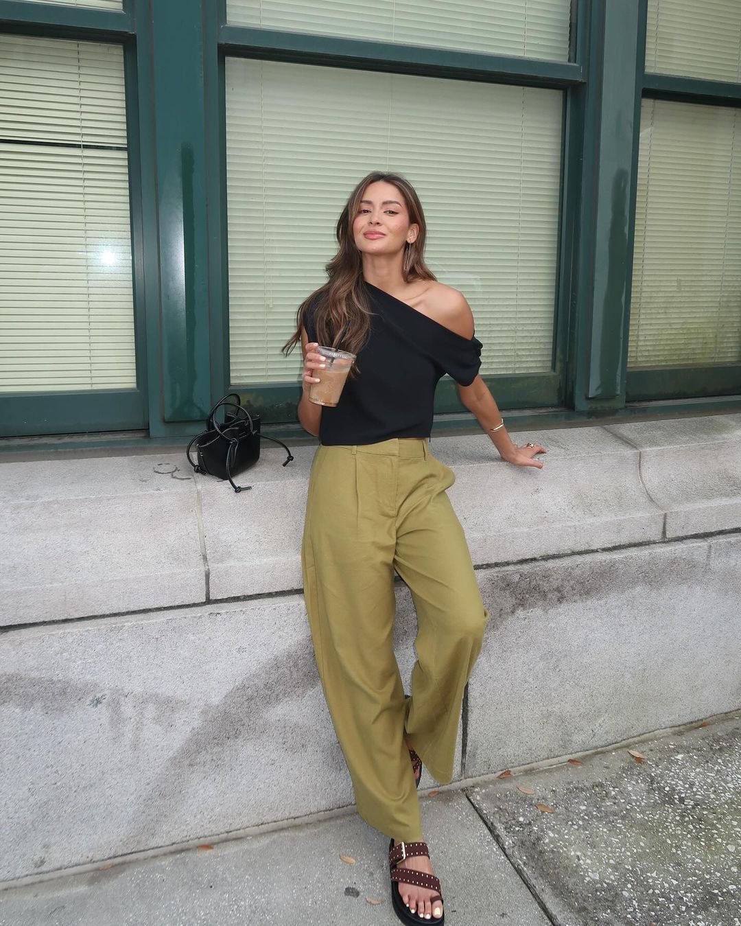 Mulher estilosa com blusa preta de ombro caído, combinada com calça wide leg verde claro e sandálias rasteiras marrons com tachas. Look casual e moderno, ideal para o clima urbano.