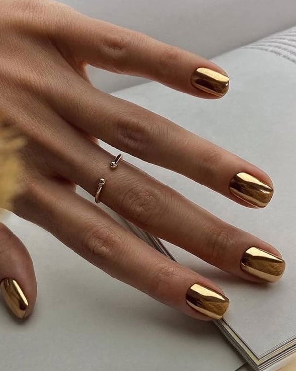 Unhas com esmalte cromado dourado destacam-se como tendência moderna, complementadas por um delicado anel minimalista.