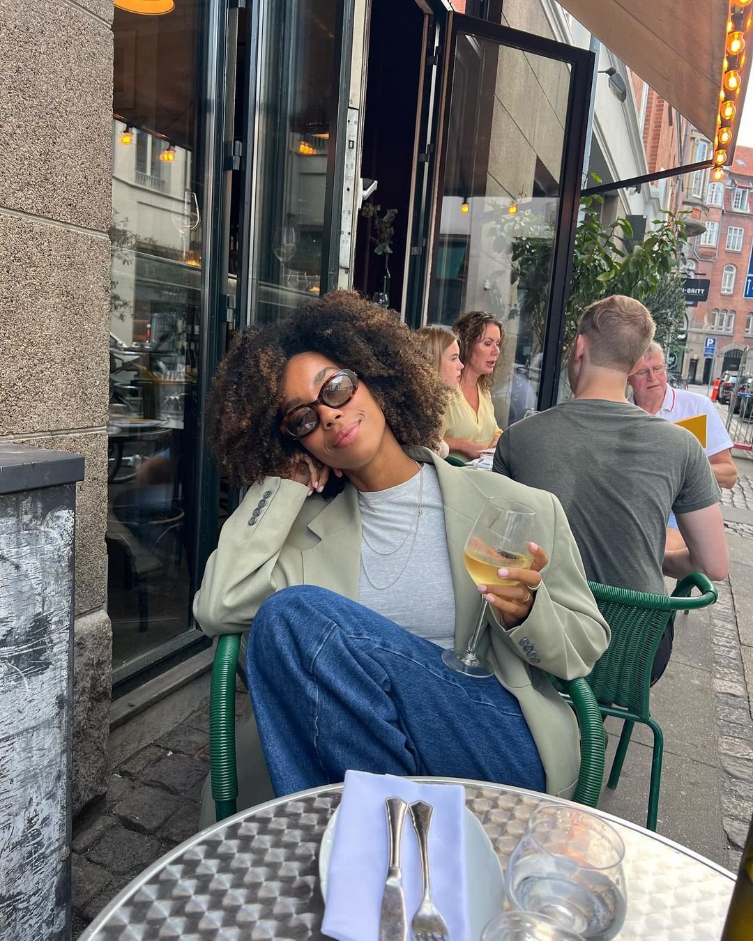 Pessoa sentada em café ao ar livre, vestindo blazer cinza sobre camiseta branca e calça jeans larga. Óculos escuros complementam o visual casual e elegante. Mulher solteira.