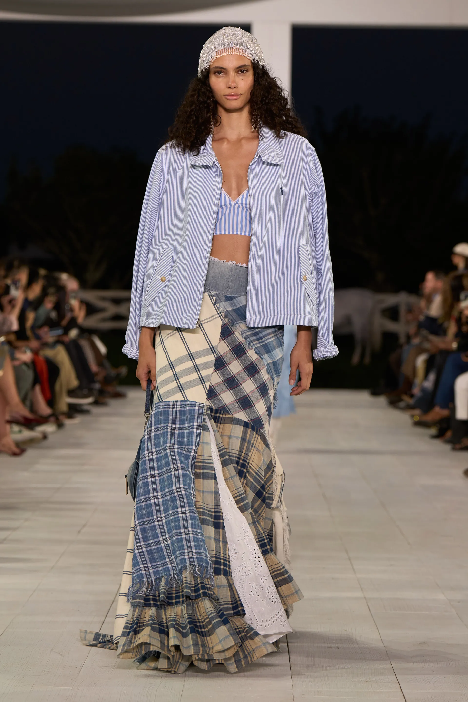 Desfile de moda: look composto por jaqueta listrada azul-clara sobre top cropped com mesma estampa. Saia longa de patchwork em tons de azul e branco, com diversas padronagens xadrez. Bandana branca com brilhos completa o visual, mesclando estilo casual e boho.