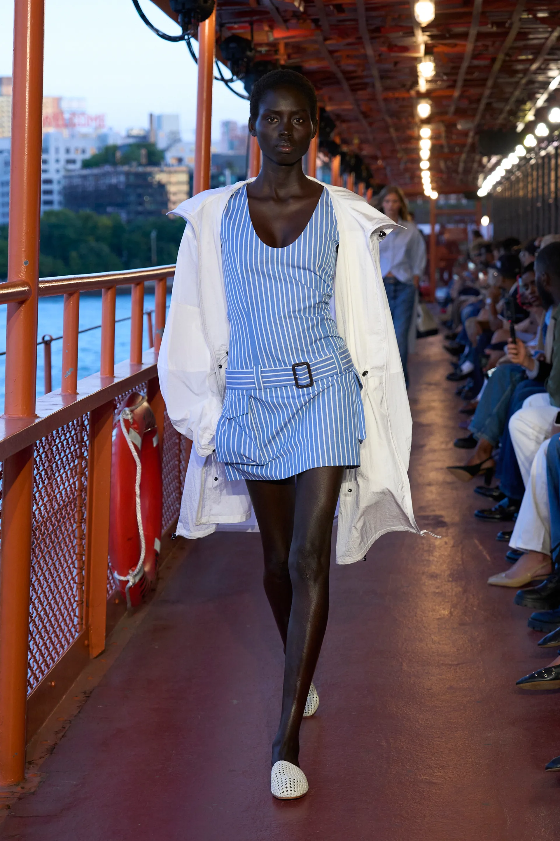 Modelo vestindo vestido curto listrado azul e branco com cinto largo e fivela. Casaco branco solto e sapatos brancos. NYFW SS25