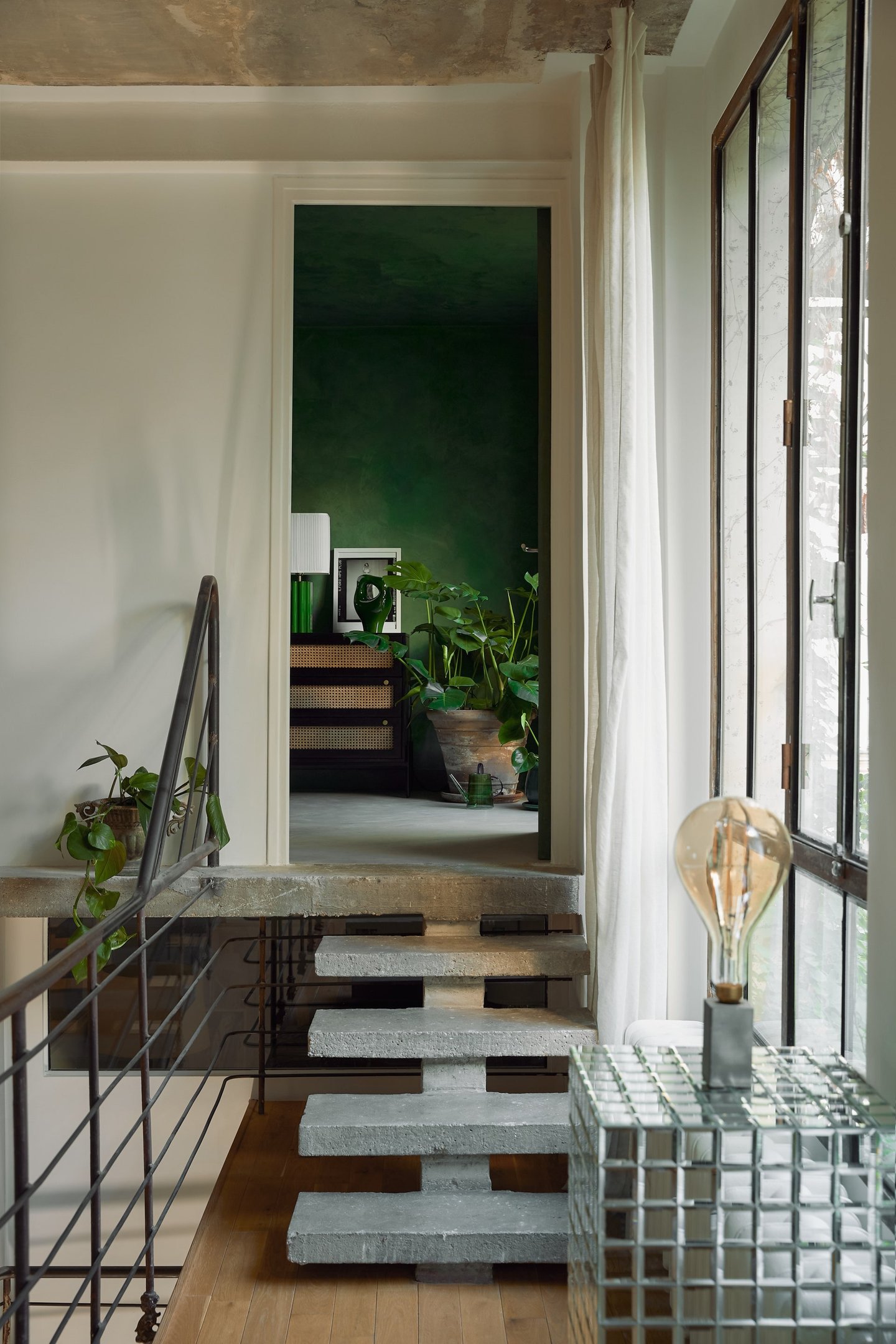 Casa moderna com decoração minimalista. Escada flutuante de concreto, paredes verde musgo e plantas internas. Ambiente iluminado por janelas amplas e luz natural, destacando a combinação de elementos industriais e naturais, tendência marcante no design de interiores.