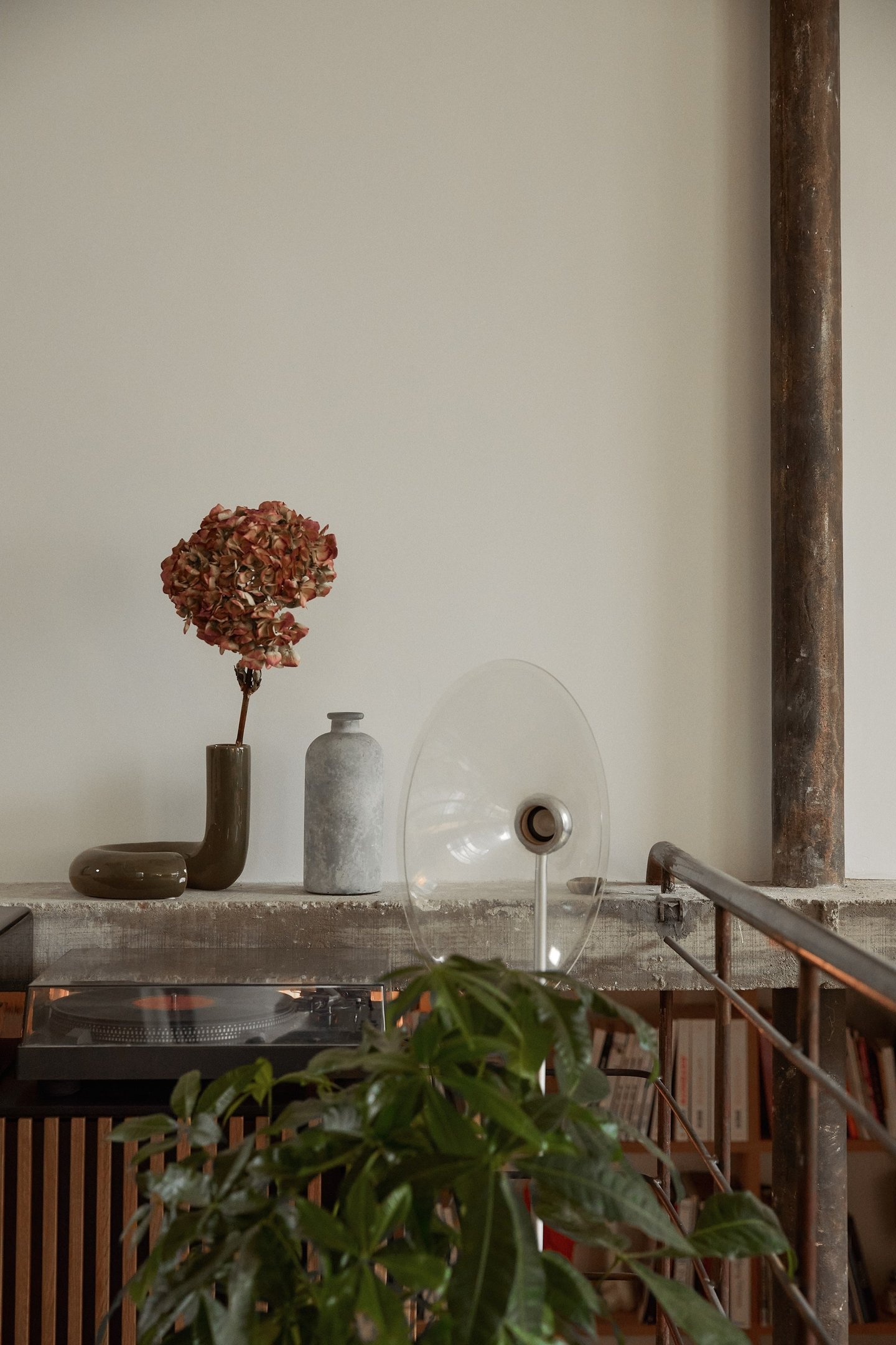 Decoração de ambiente interno com tendências minimalistas e rústicas. Objetos decorativos incluem um vaso com flores secas em tom terroso, uma garrafa de cerâmica cinza e uma planta verde. Ambientes de estilo industrial com uso de metal e madeira.