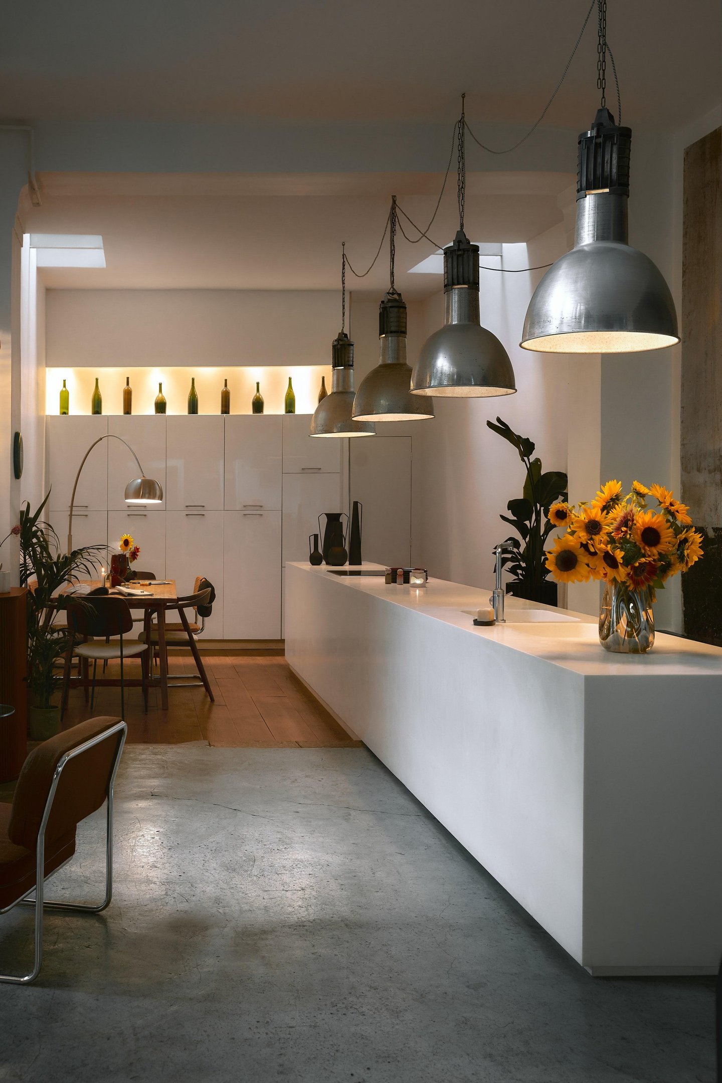 Cozinha moderna com estética minimalista. Ilha branca com bancada clean, luminárias industriais suspensas e decoração com girassóis. Ambiente iluminado com luz natural e garrafas decorativas. Estilo contemporâneo com uso de madeira e metais.