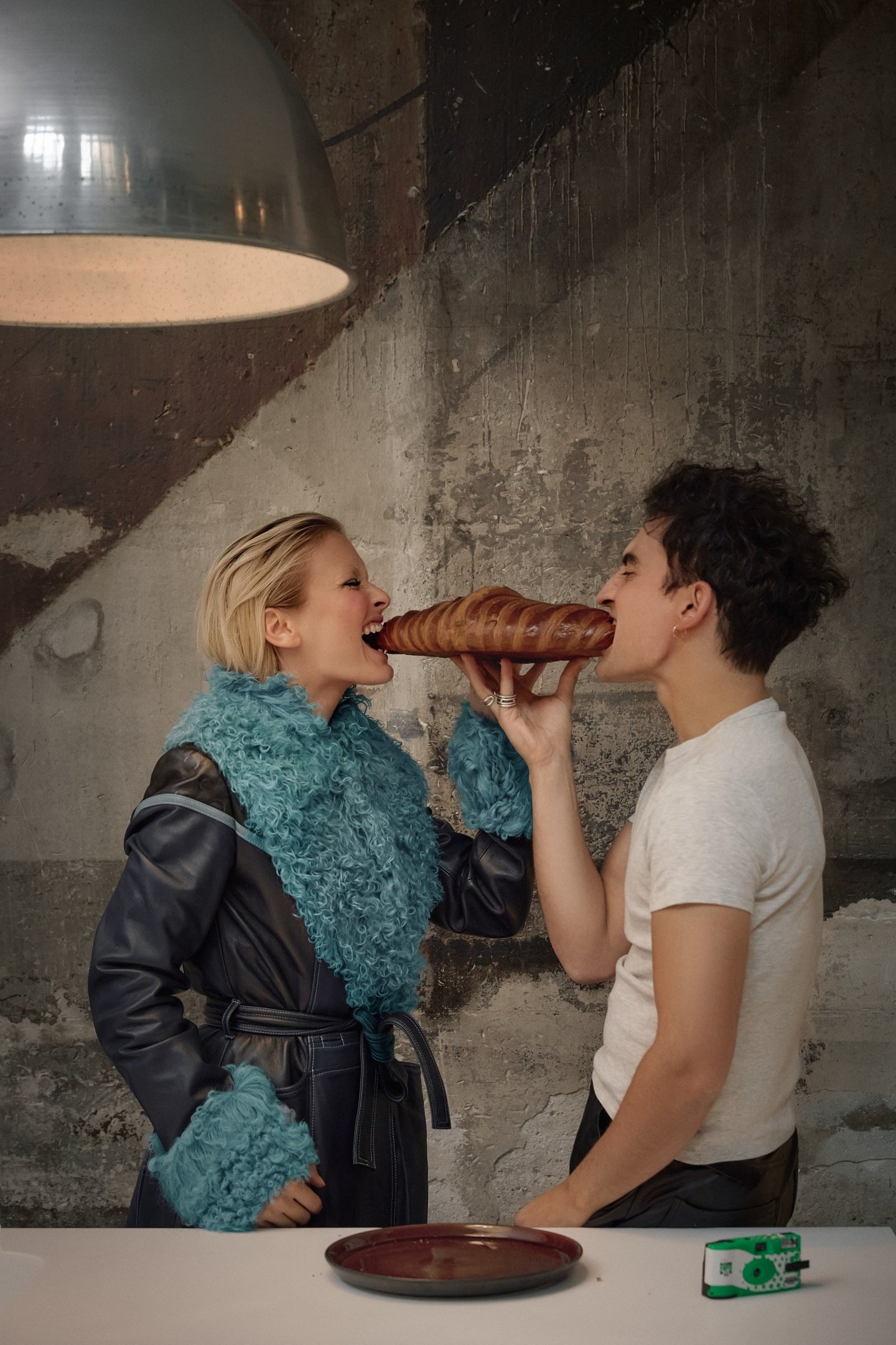 Imagem de duas pessoas com estilos contrastantes de moda. A mulher usa um casaco azul com detalhes de pele fake, destacando uma tendência moderna e ousada. O homem veste uma camiseta branca básica, representando um look casual e descontraído. Eles dividem um pão grande, adicionando um toque de humor.